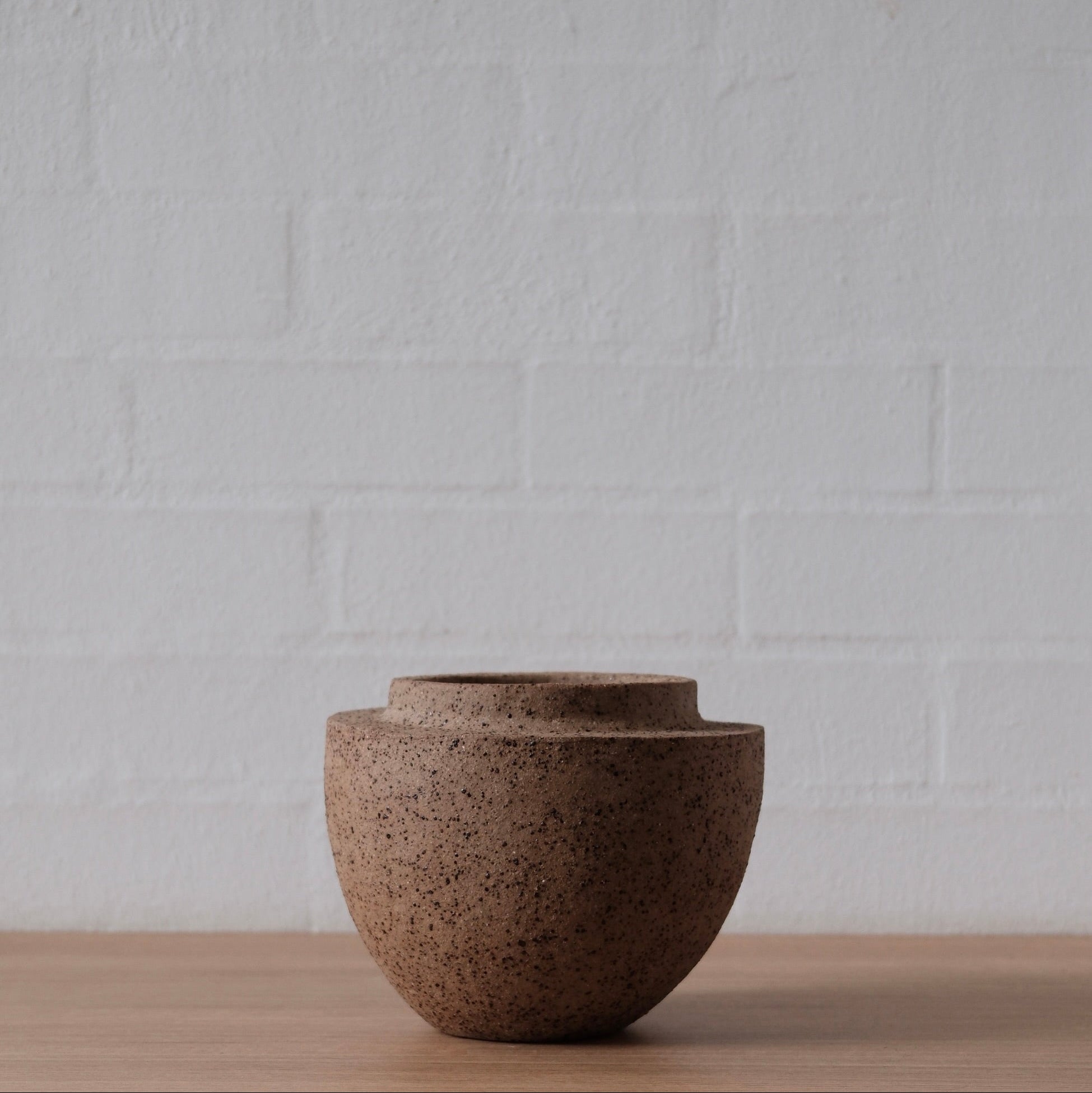 Warm brown, speckled ceramic vessel on a wooden surface with a white textured wall background