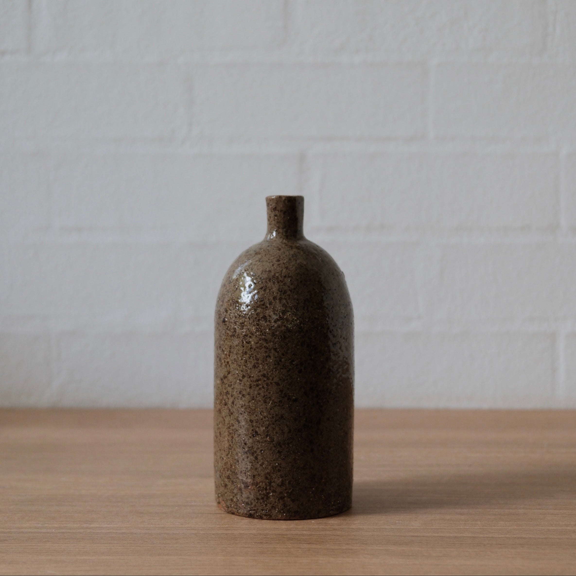 Medium olive speckled ceramic vase, with cylindrical body, rounded top and small spout, one a wooden bench in front of a white wall