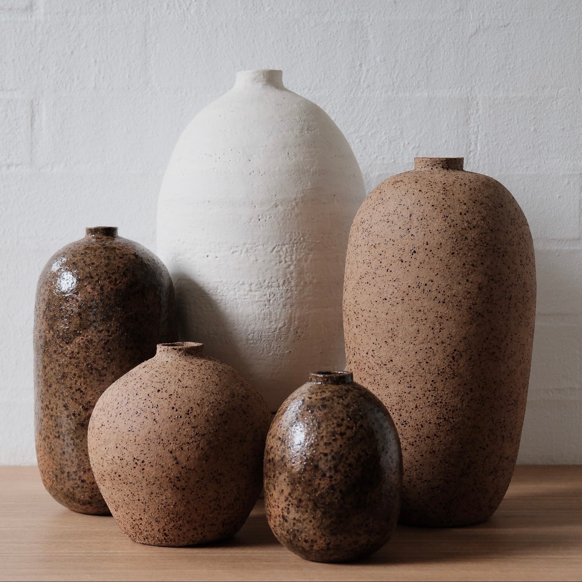 Set of ceramic vessels with textured surfaces ranging from white to wam speckled brown on a wooden surface against a white wall.