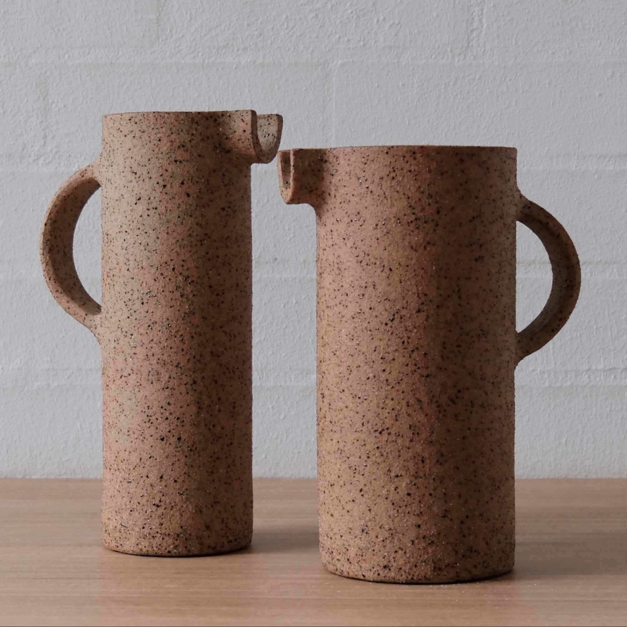 Two tall cylindrical ceramic pitchers with speckled brown matt finish, displayed on a light wood surface against a white brick wall.