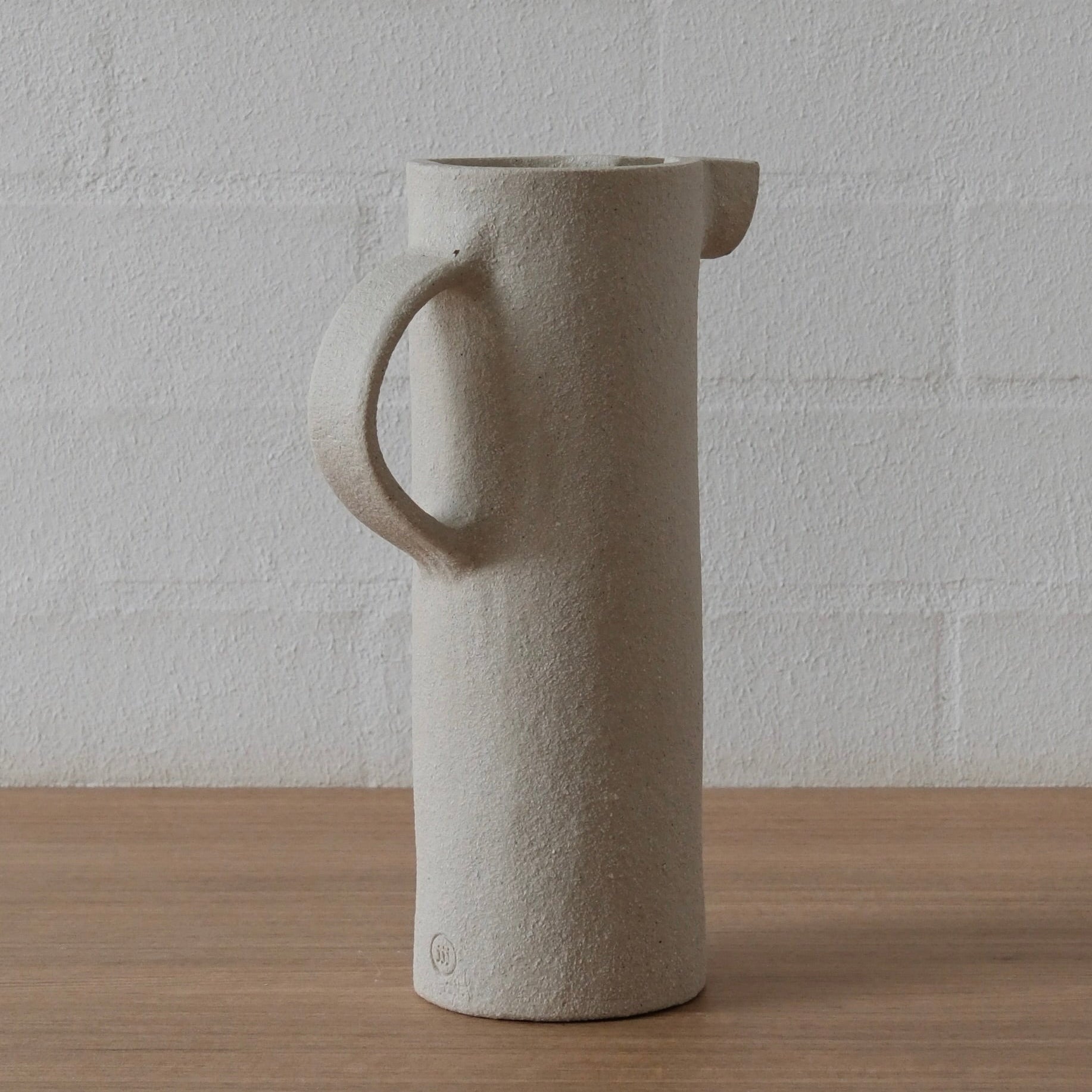 A tall, cylindrical ceramic pitcher with a textured white matte finish, featuring a loop handle on the left and a small pouring spout on the right, set on a wooden surface against a white brick wall.
