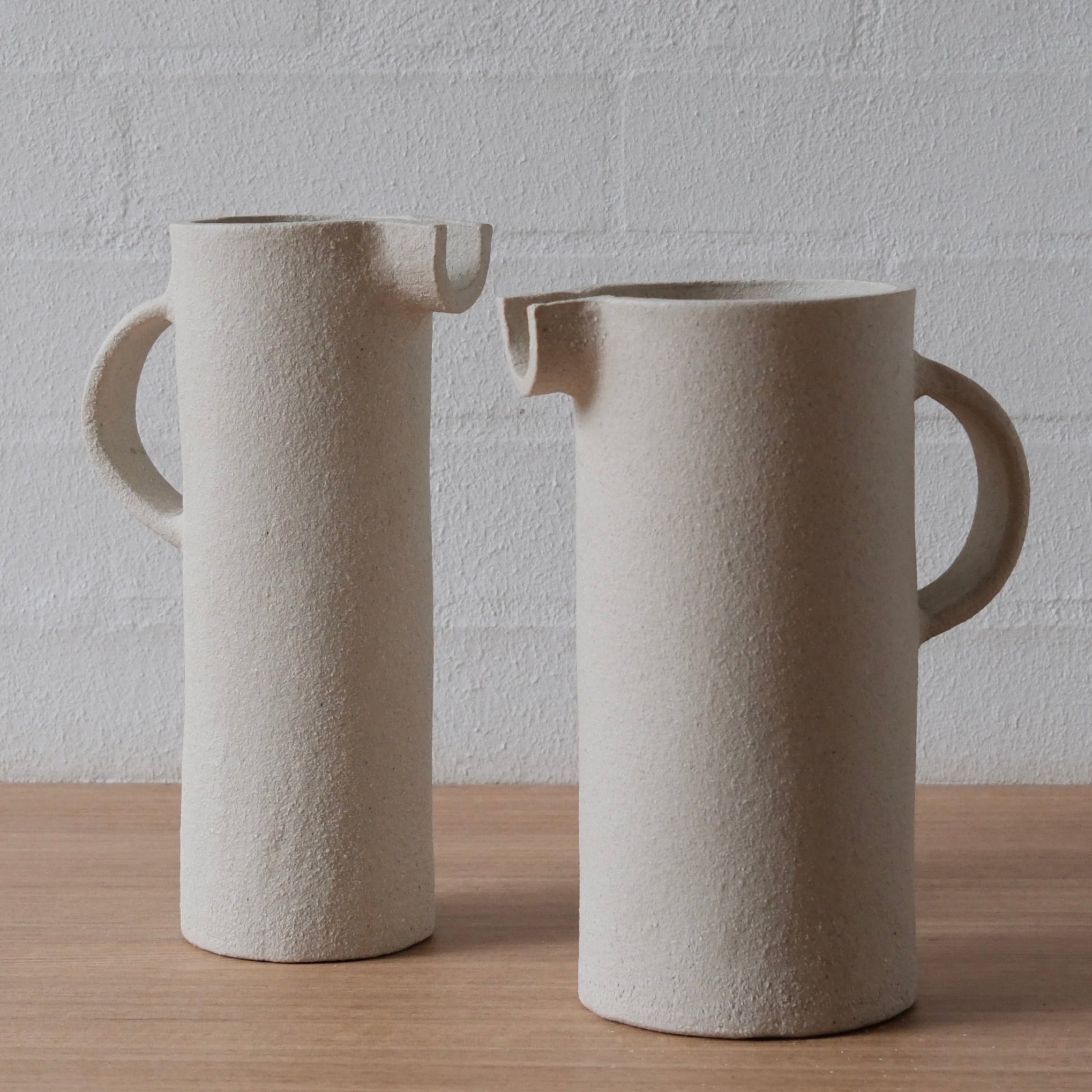 Two white matte ceramic pitchers with textured surfaces, shown on a light wood surface against a white brick wall background.