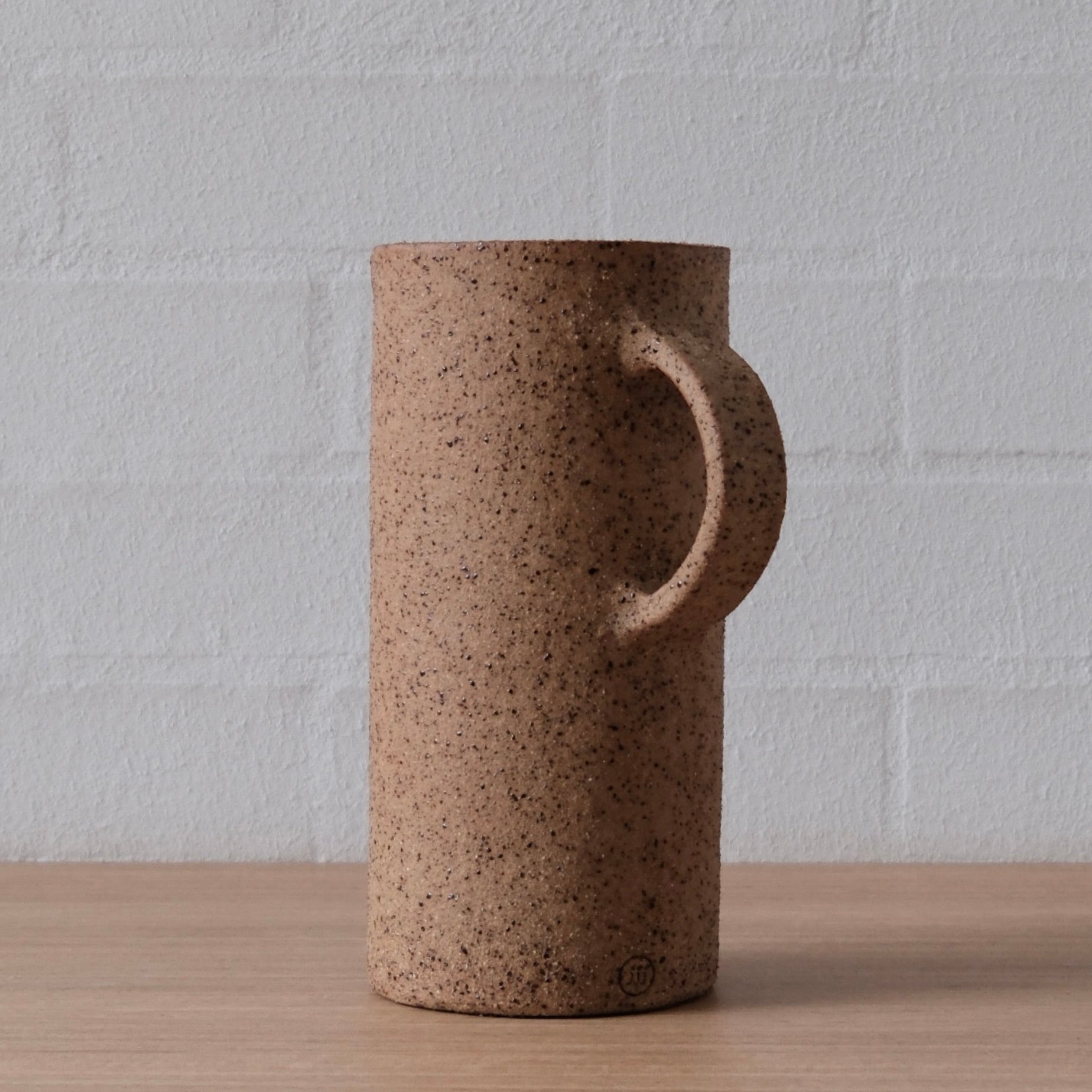 A cylindrical ceramic pitcher with a textured, speckled brown finish, featuring a unique integrated handle, set on a light wood surface against a white brick wall background.