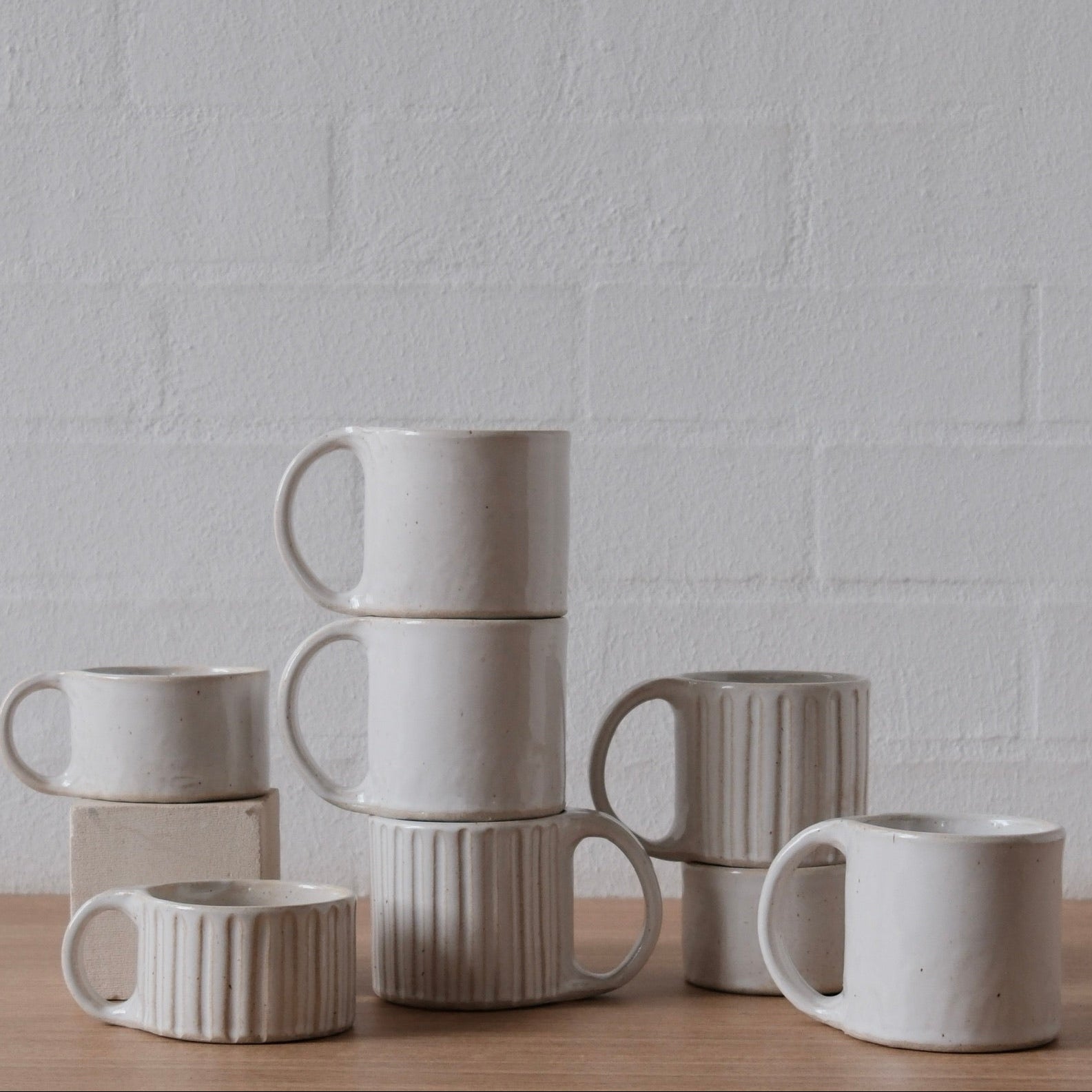 A selection of small and regular, ribbed and plain, white ceramic mugs, arranged in stacks and pairs on a light wood surface against a white textured brick wall.