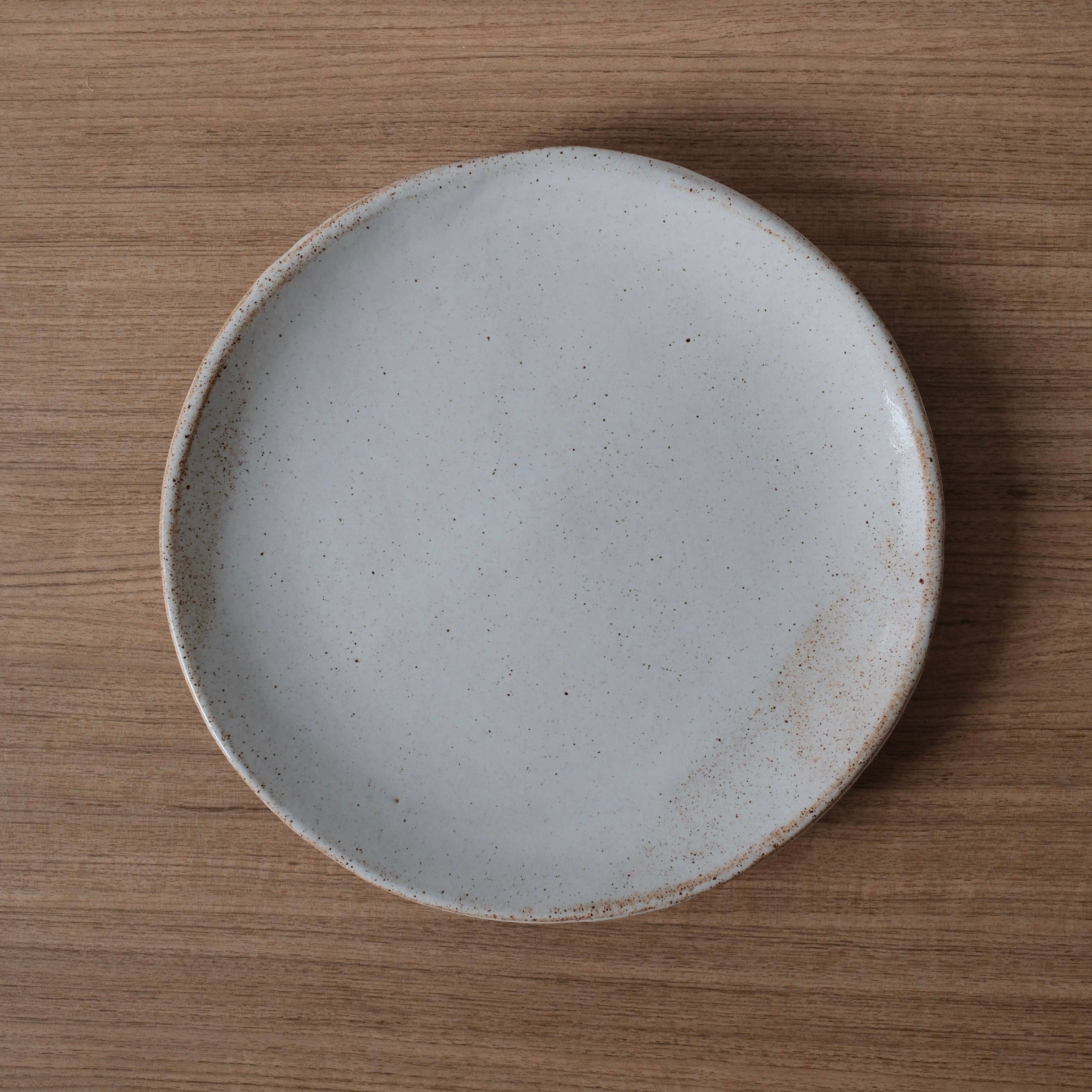 Round white ceramic dinner plate with a slightly speckled finish and golden highlights on a wooden surface