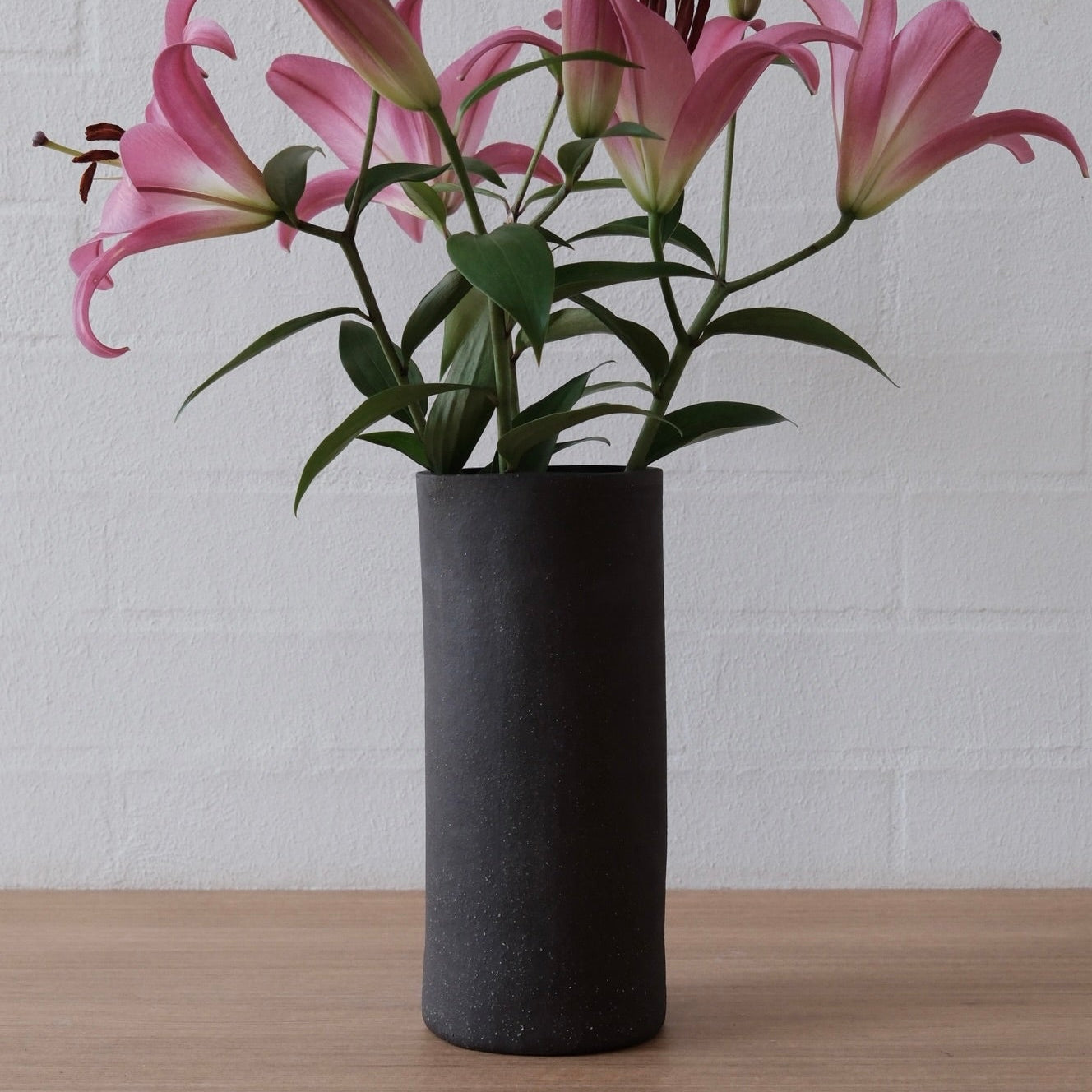 Pink lilies arranged in a tall matte black cylindrical vase on a light wood surface against a white brick wall.