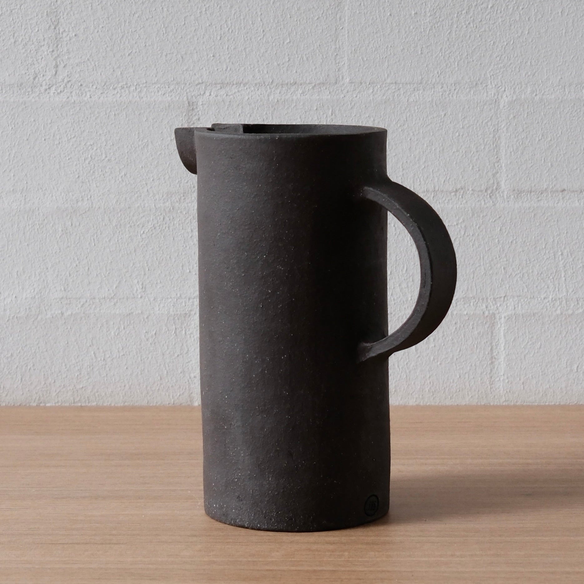 A matte black ceramic pitcher with a large integrated handle and a small pouring spout, standing on a light wood surface against a white brick wall.
