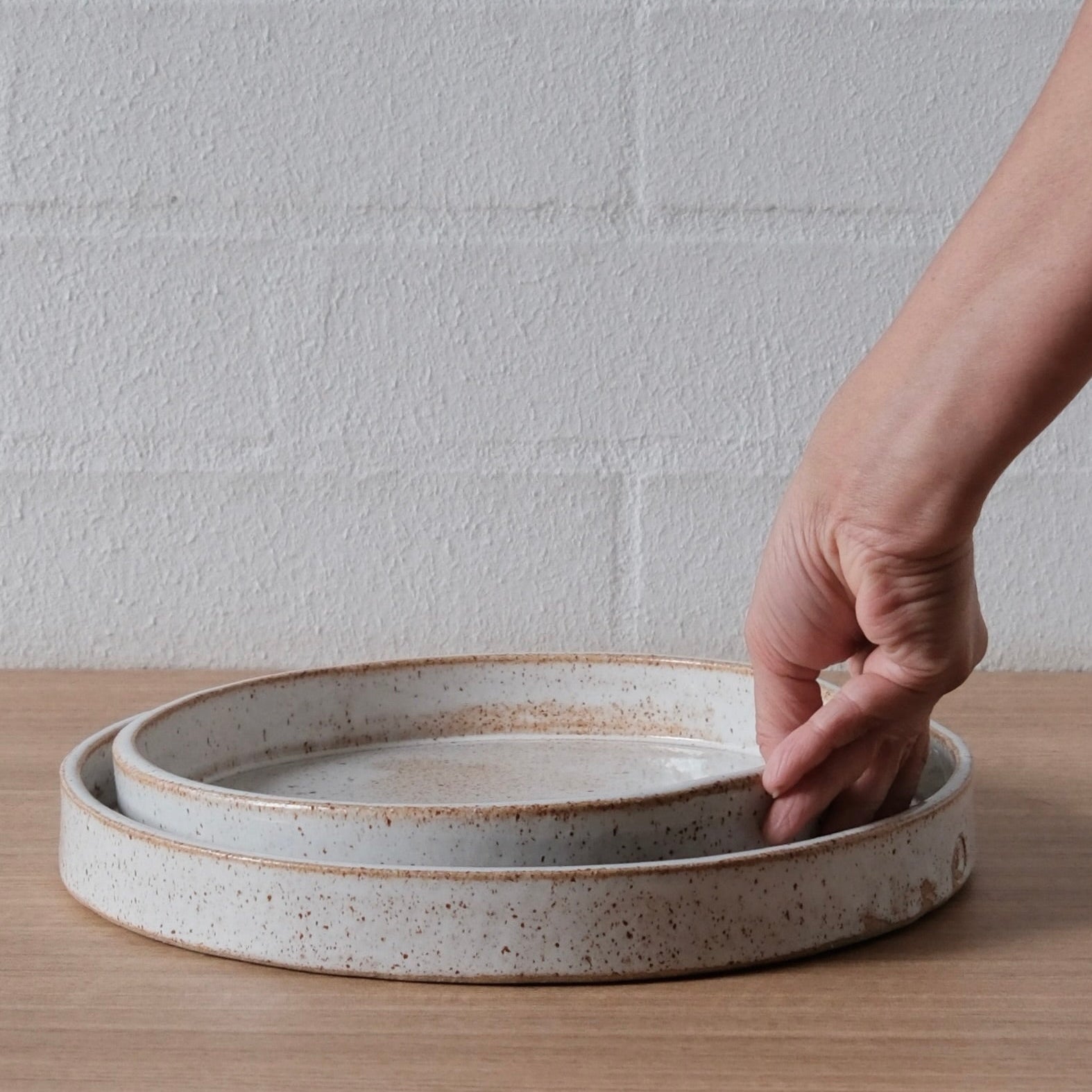 A hand is shown lifting the edge of a round ceramic tray, which sits on top of two nested shallow ceramic plates with a speckled off-white glaze and brown rim detailing, set on a wooden surface against a white brick wall.