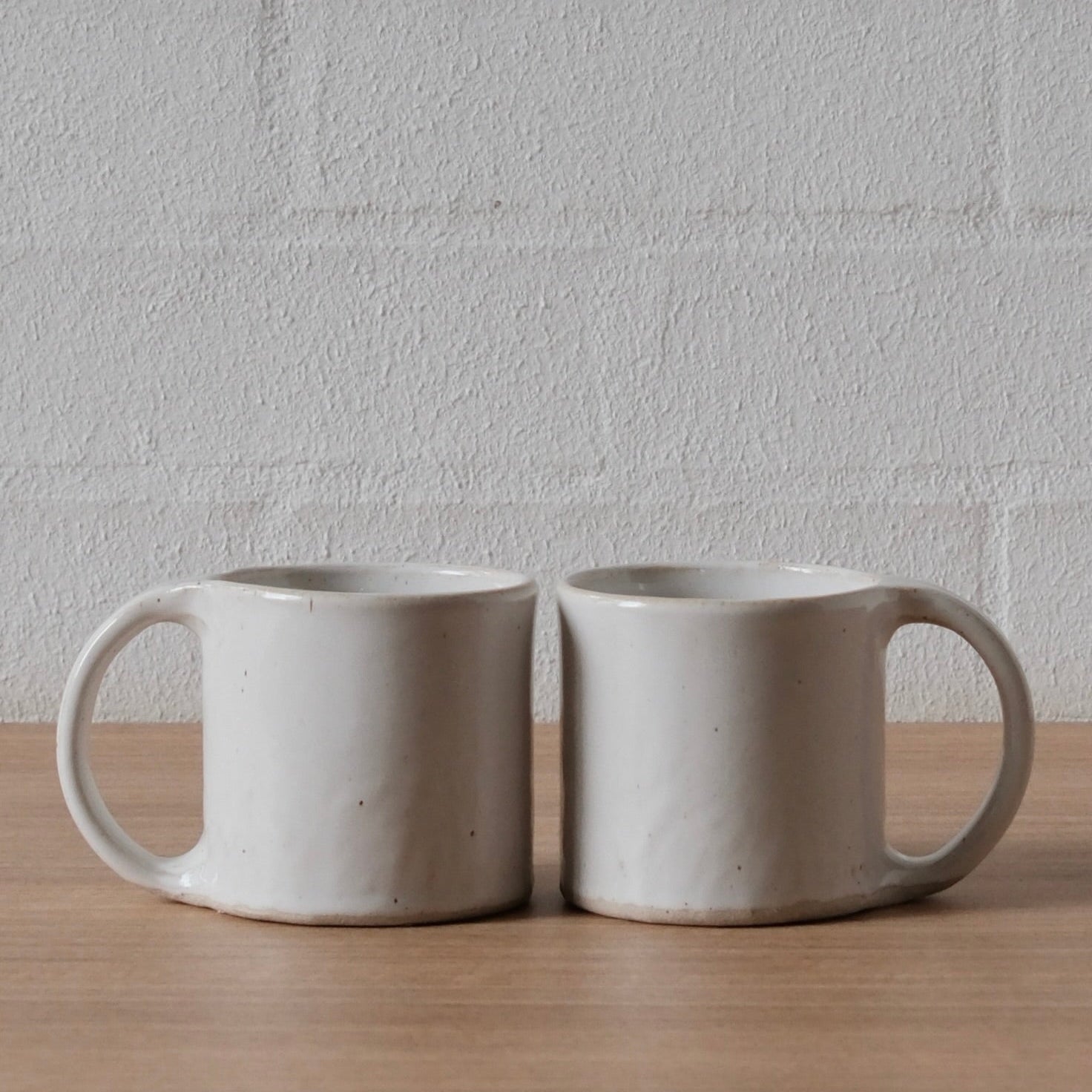 Two handmade white ceramic mugs sitting side-by-side on a light wood surface against a white brick wall.