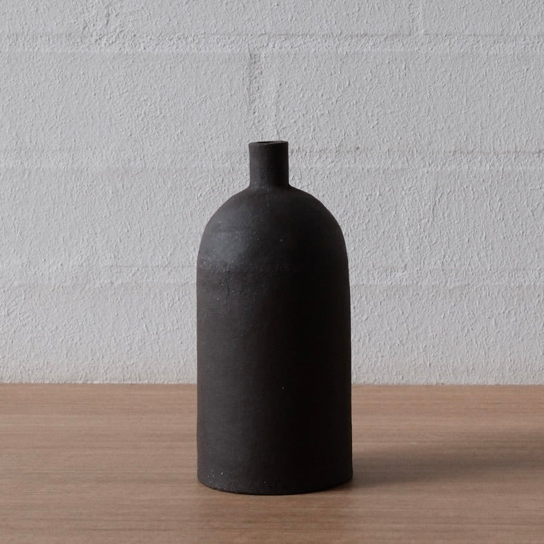 A matte black ceramic bottle with a short, cylindrical neck and a rounded body, standing on a light wood surface against a white painted brick wall.