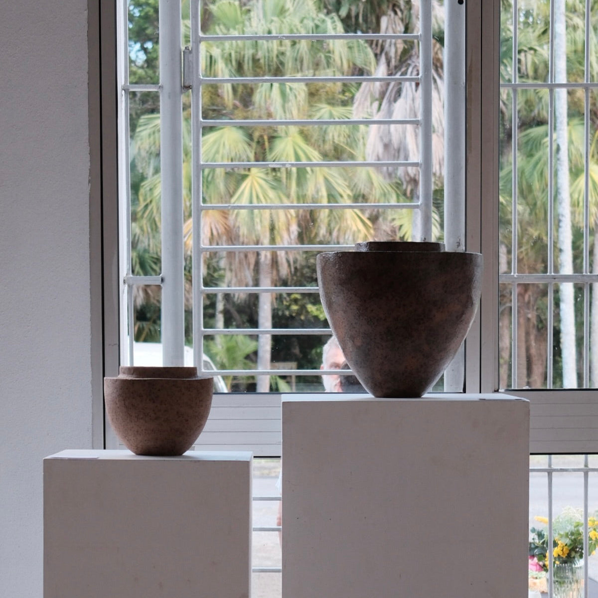 Two brown ceramic vessels on white plinths with a window view of trees.