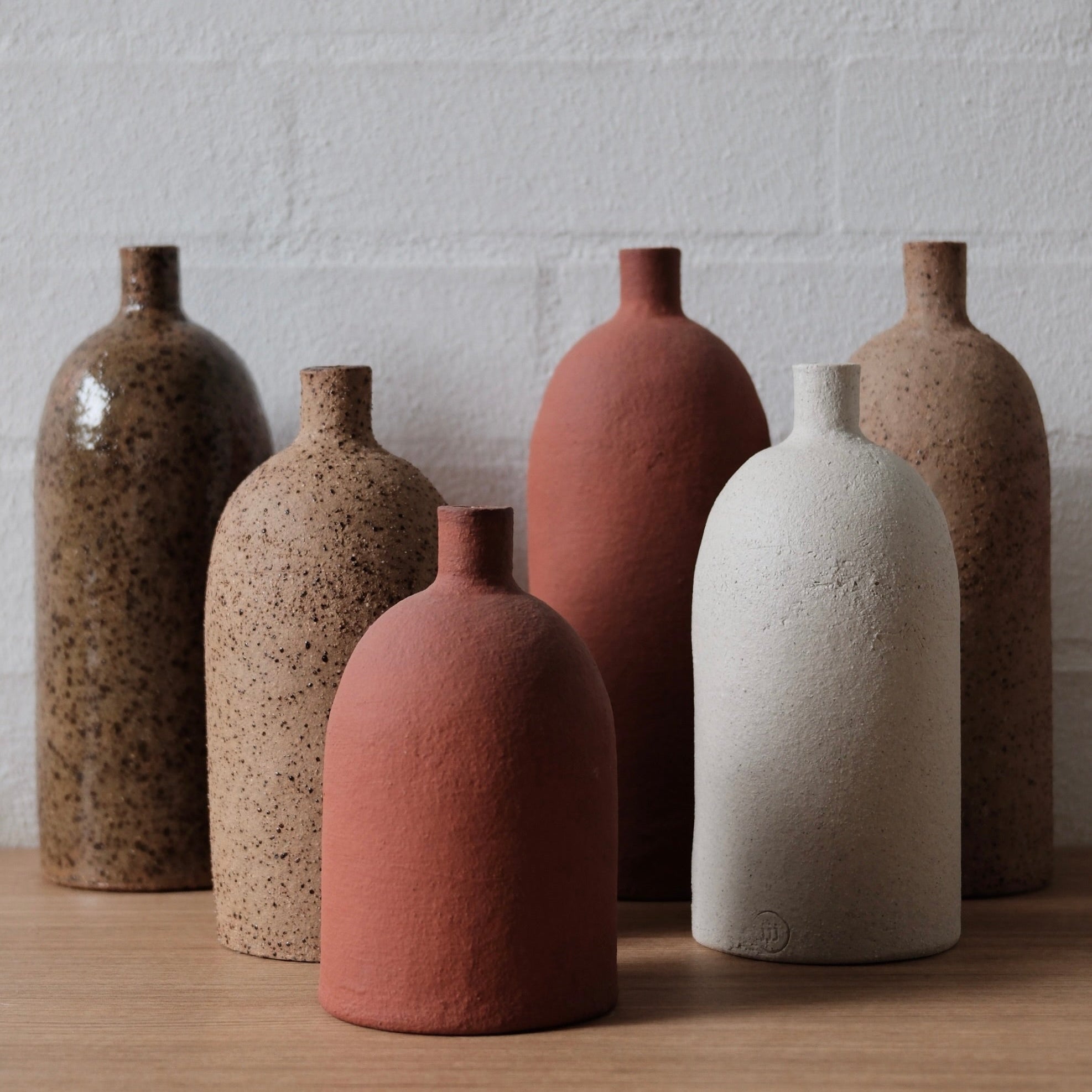 Set of ceramic vases, with cylindrical body, rounded top and small spout, in terracotta, white and speckled brown, on a wooden bench in front of a white wall