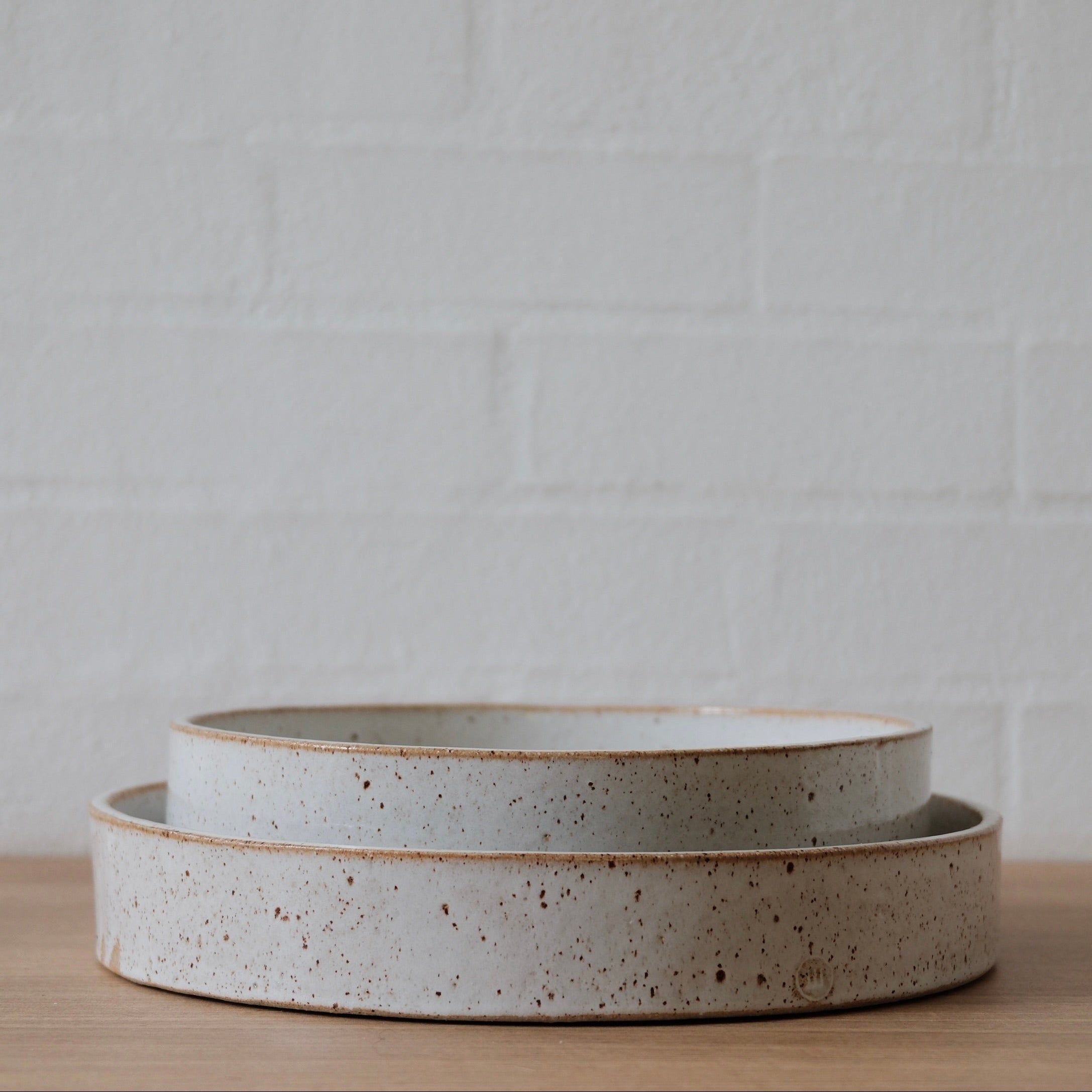 Two white ceramic bowls with straight sides stacked one inside the other, with slightly speckled golden finish on a wooden surface against a white textured wall.