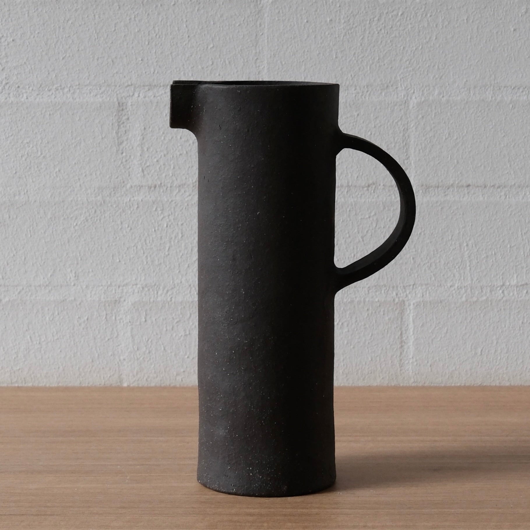 A matte black ceramic pitcher with a cylindrical body, a loop handle, and a pouring spout, standing on a light wood surface against a white brick wall background.