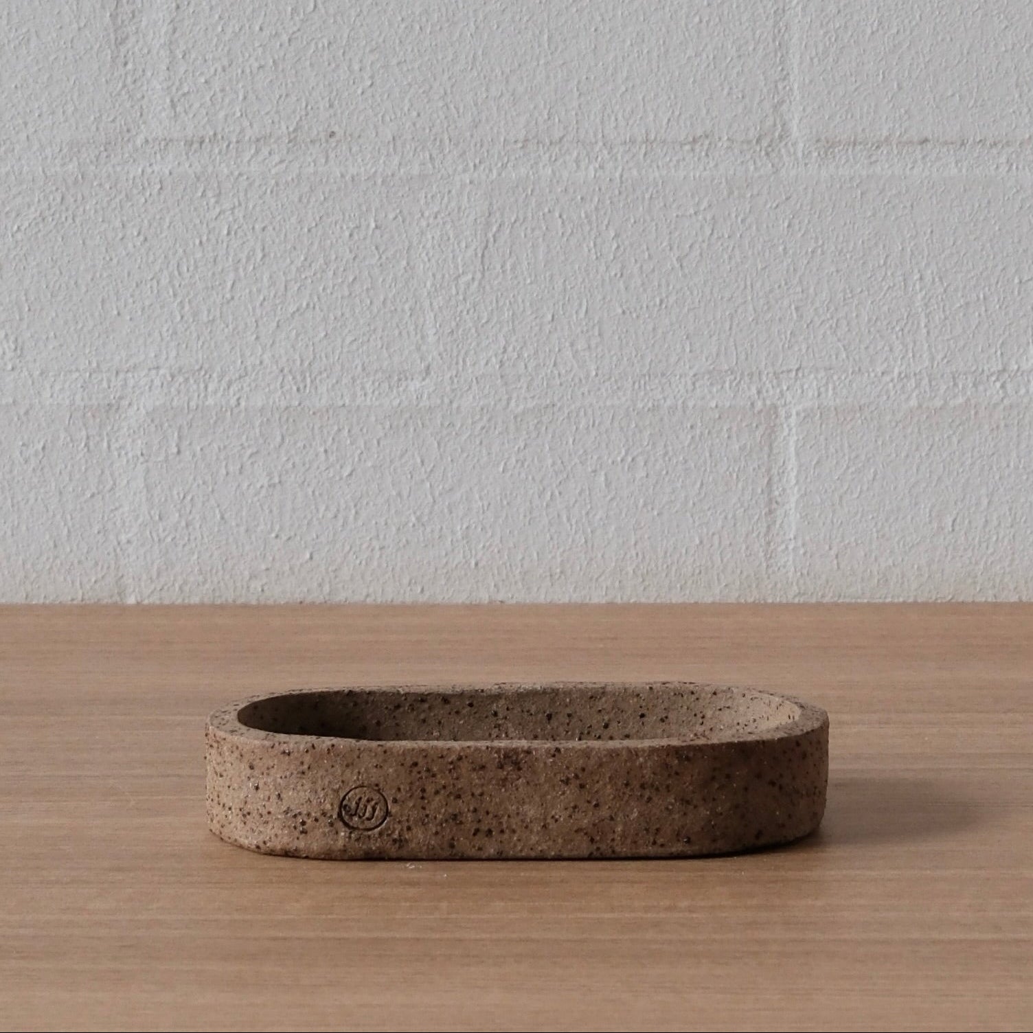 A small oval ceramic dish with a textured, speckled brown finish, sitting on a light wood surface against a white brick wall background.
