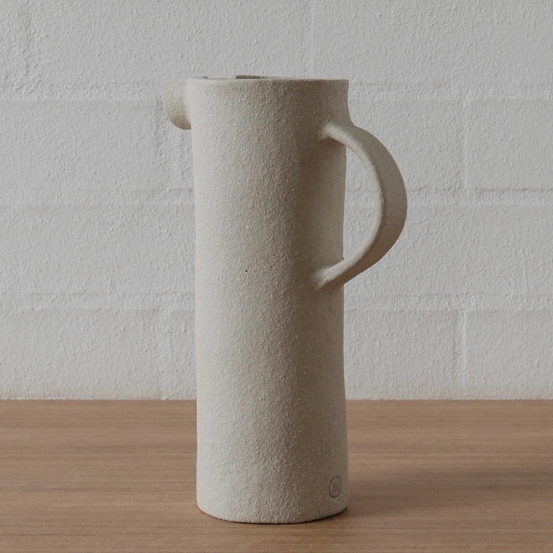 A tall, cylindrical ceramic pitcher with a textured, white matte finish, featuring a curved handle and a small pouring spout, set on a light wood surface against a white brick wall.