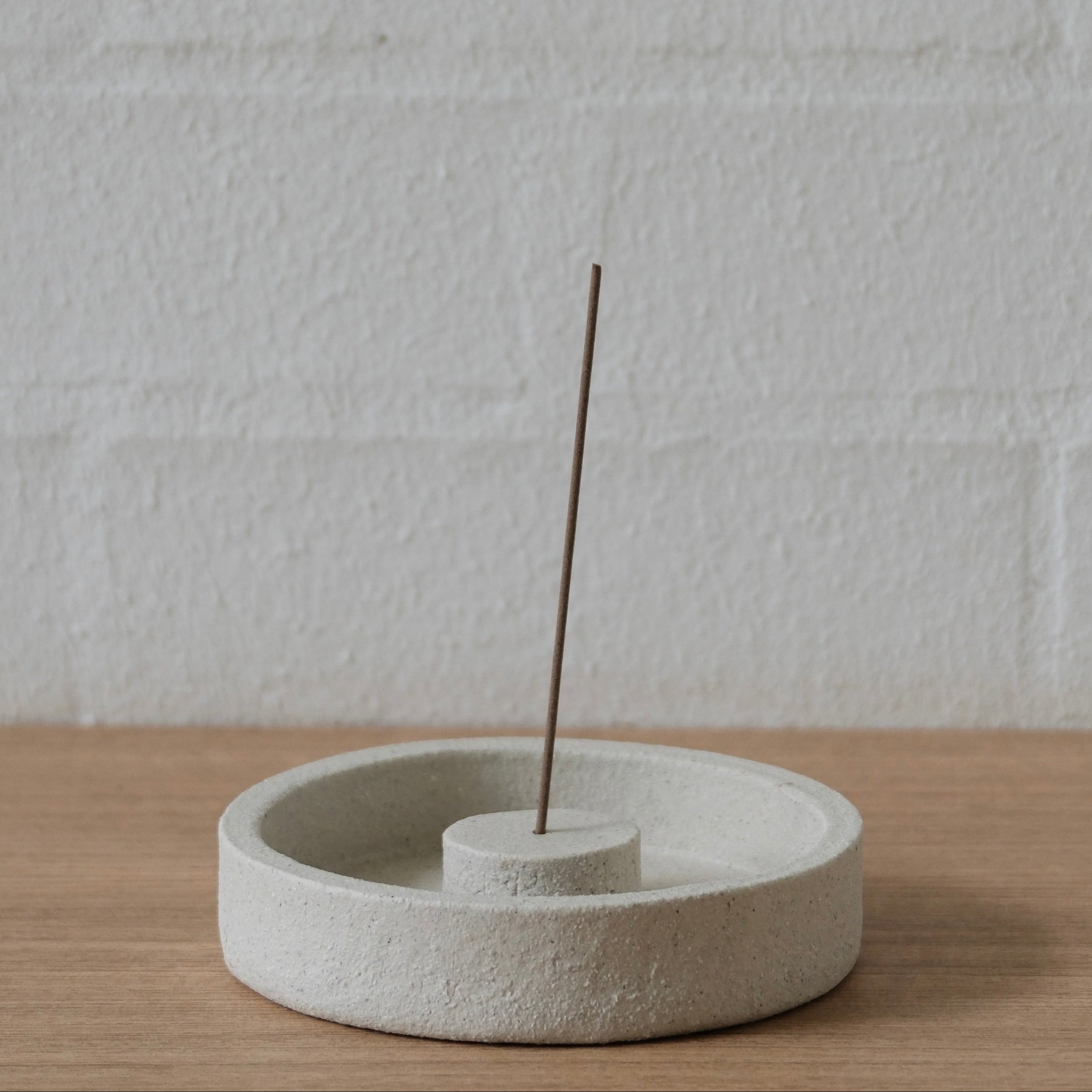 Textured, white, round ceramics incense holder with a single incense stick on a light wooden surface and white wall background.