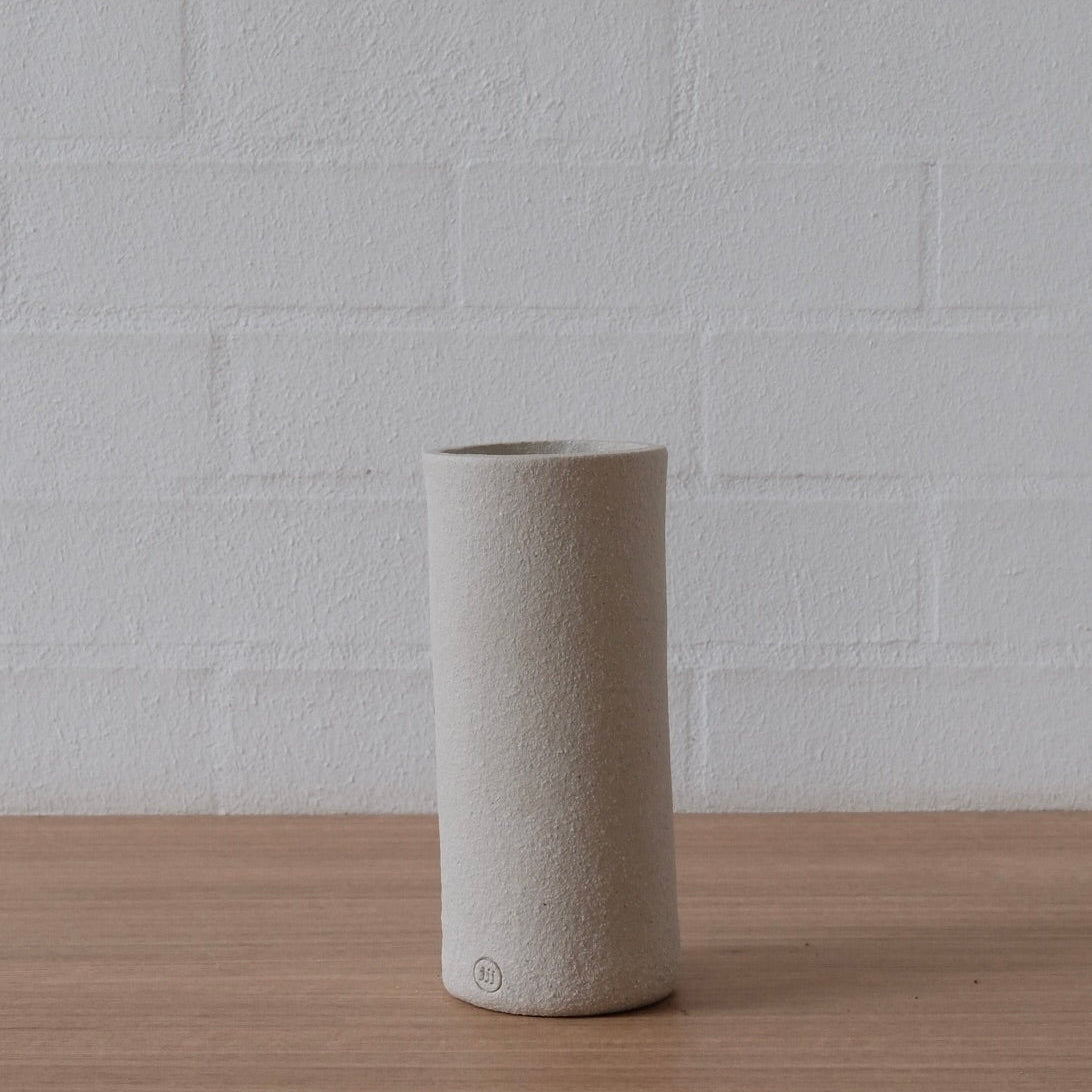 A minimalist, cylindrical ceramic vase in a textured white finish, standing on a light wood surface against a white brick wall background.