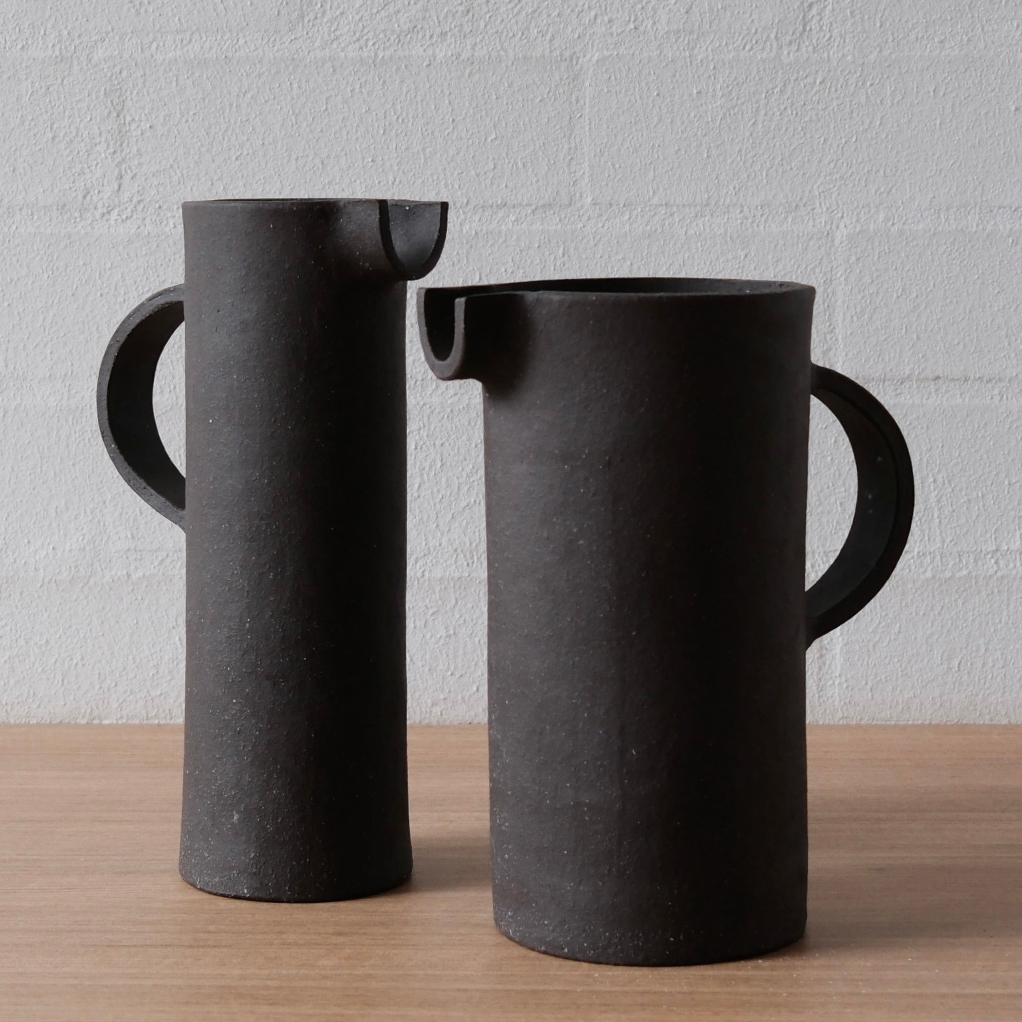 Two matte black ceramic pitchers with integrated handles and small pouring spouts, standing on a light wood surface against a white painted brick wall.
