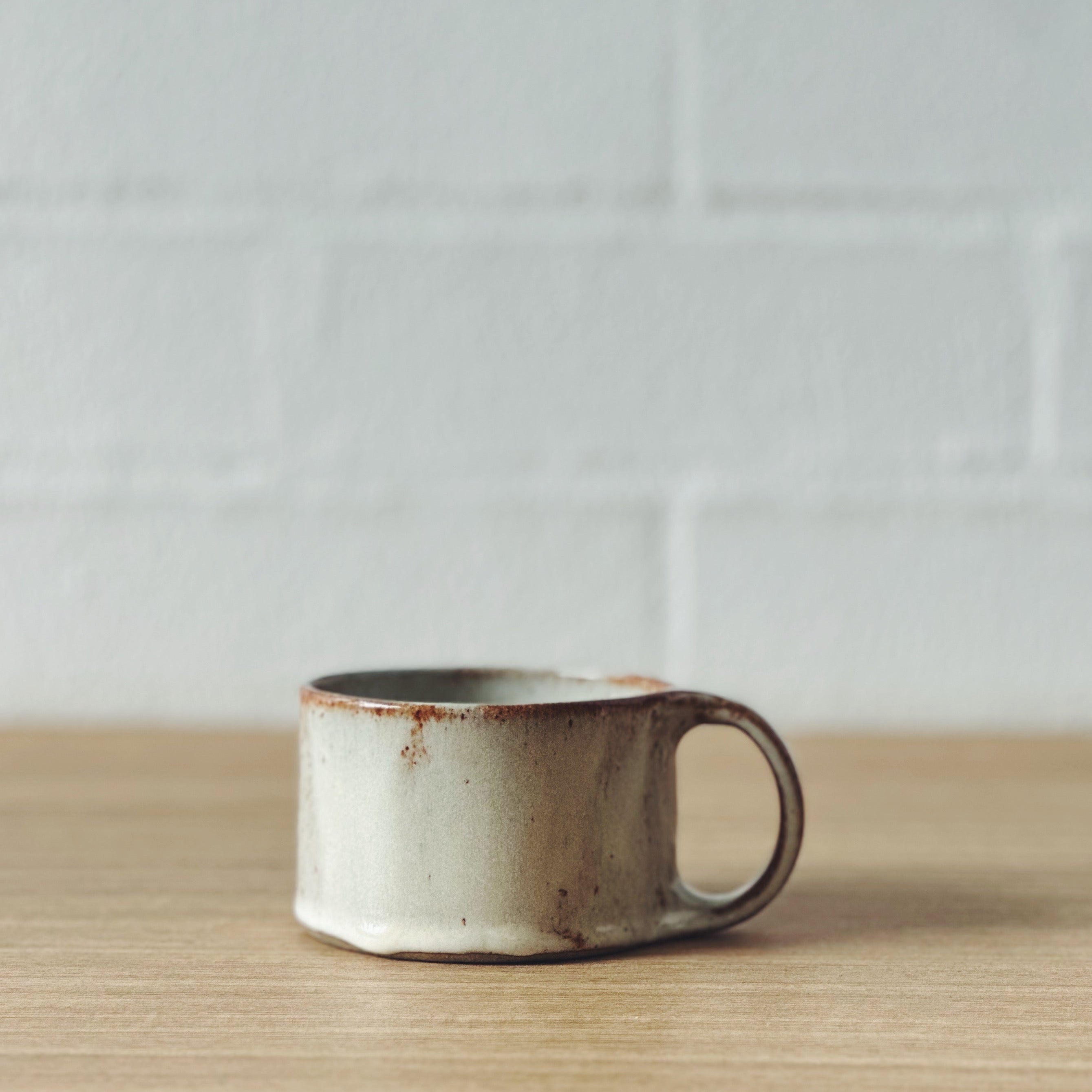 A small handmade ceramic cup with a rustic finish, featuring a light beige body with brown speckles and a dark brown rim, sitting on a light wood surface against a white brick wall background.
