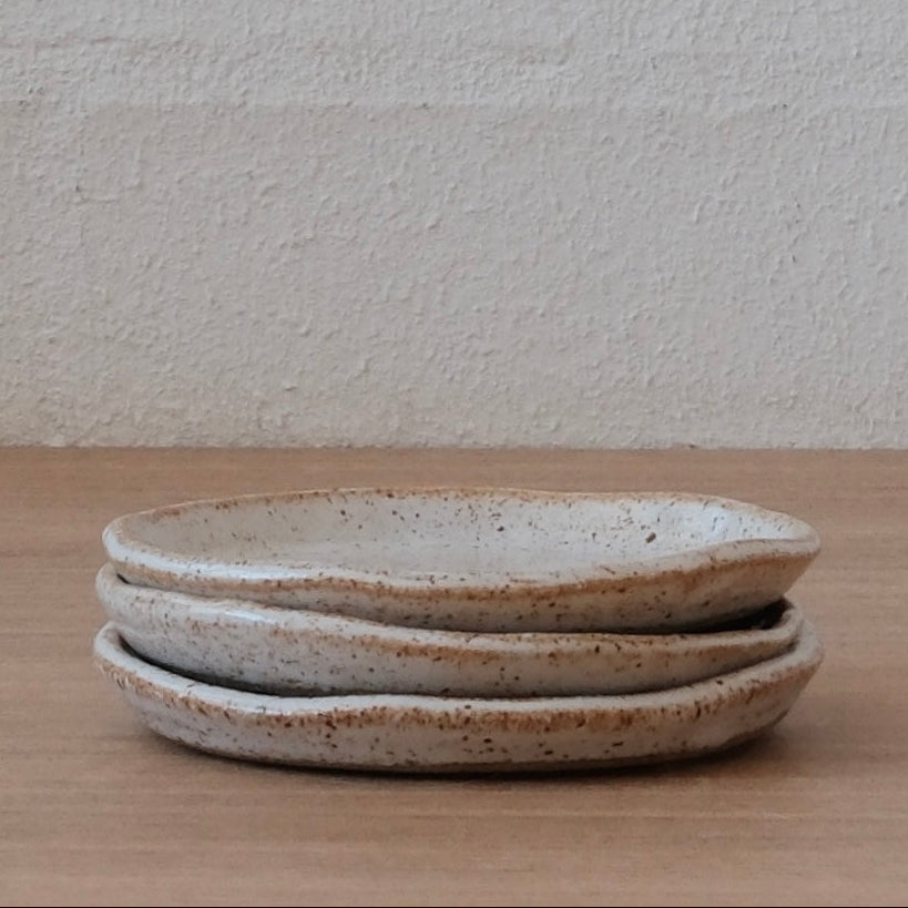 Side view of a stack of three small white ceramic plates with slightly speckled finish and golden highlights on a wooden surface and textured white background