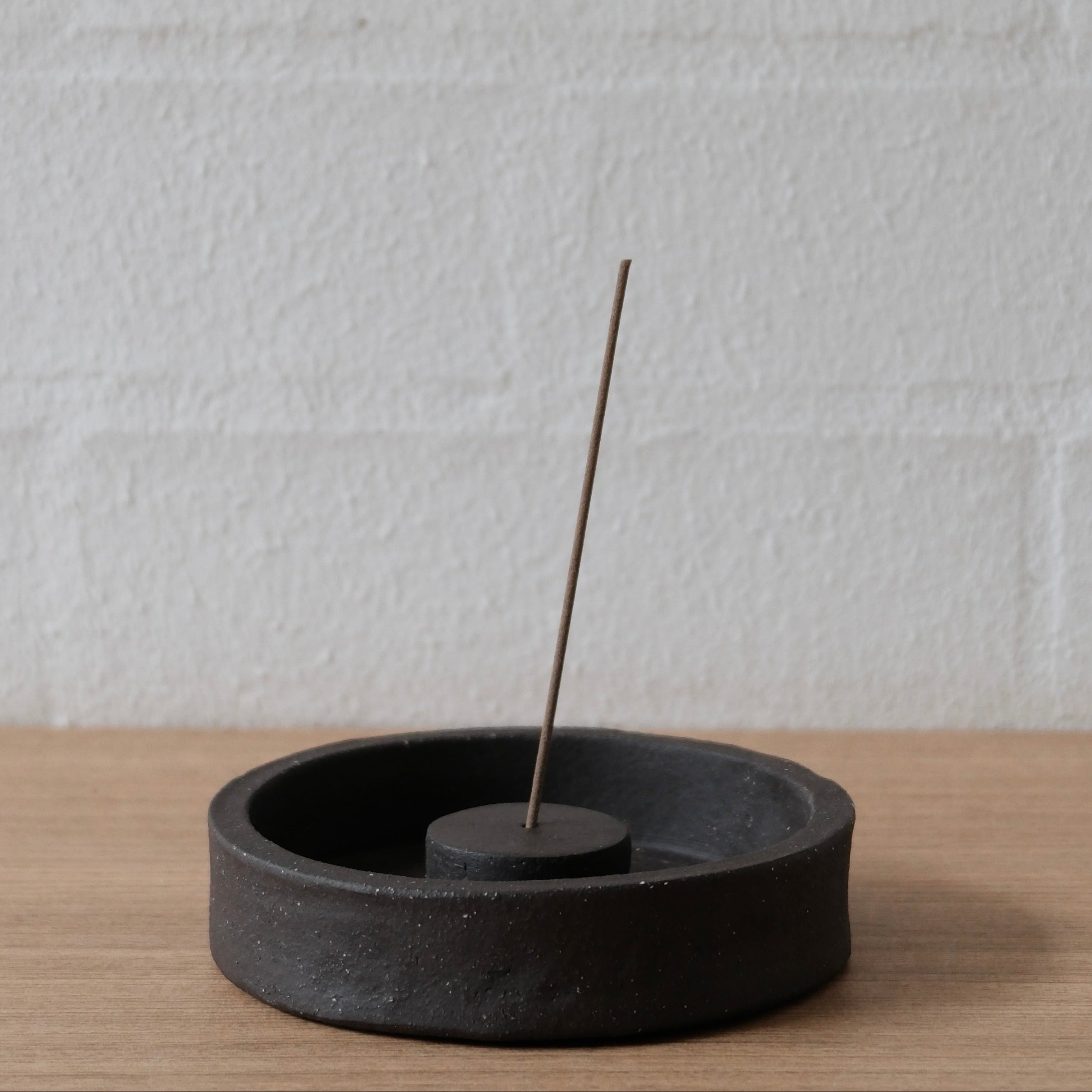 Black ceramic incense holder with a single incense stick on a light wooden surface and white textured wall background.