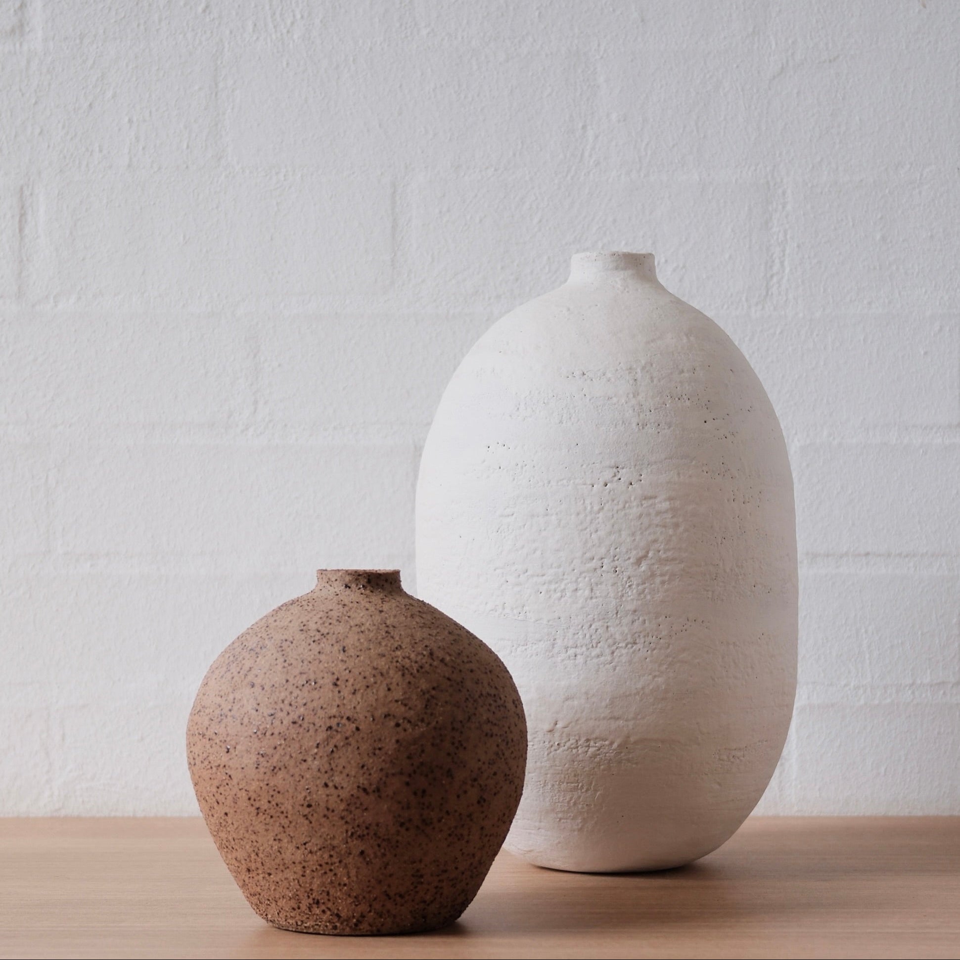 Two ceramic vases, one brown and one white, on a wooden surface with a white brick wall background.