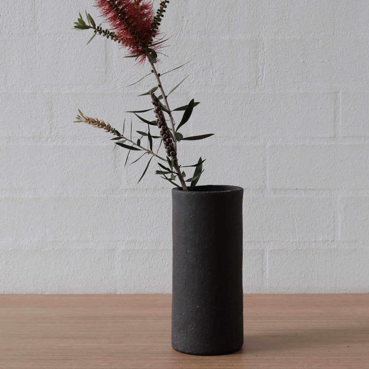 A matte black cylindrical ceramic vase holding a dried botanical arrangement with green stems and a dusty burgundy flower, set on a light wood surface against a white painted brick wall.