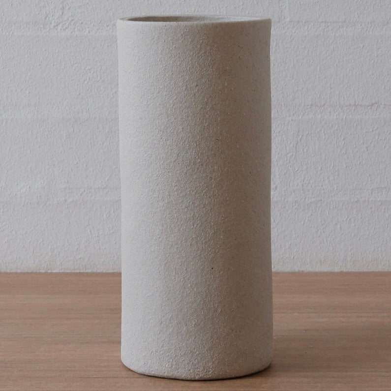 A tall, cylindrical ceramic vase with a textured, white matte finish, standing on a light wood surface against a white brick wall background.