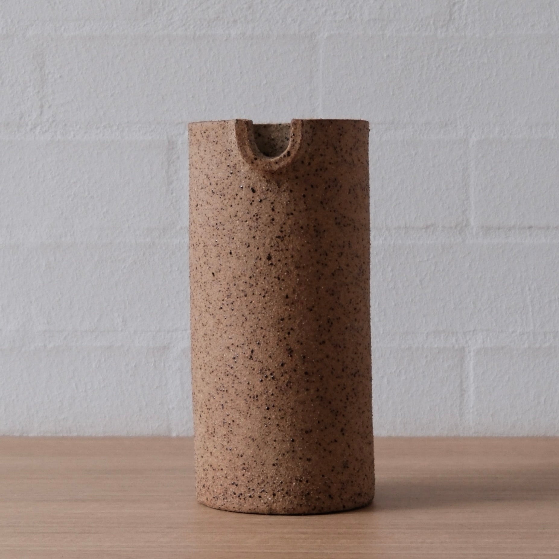 A cylindrical ceramic vase with a textured, speckled brown finish and a small cutout at the top, standing on a light wood surface against a white brick wall background.