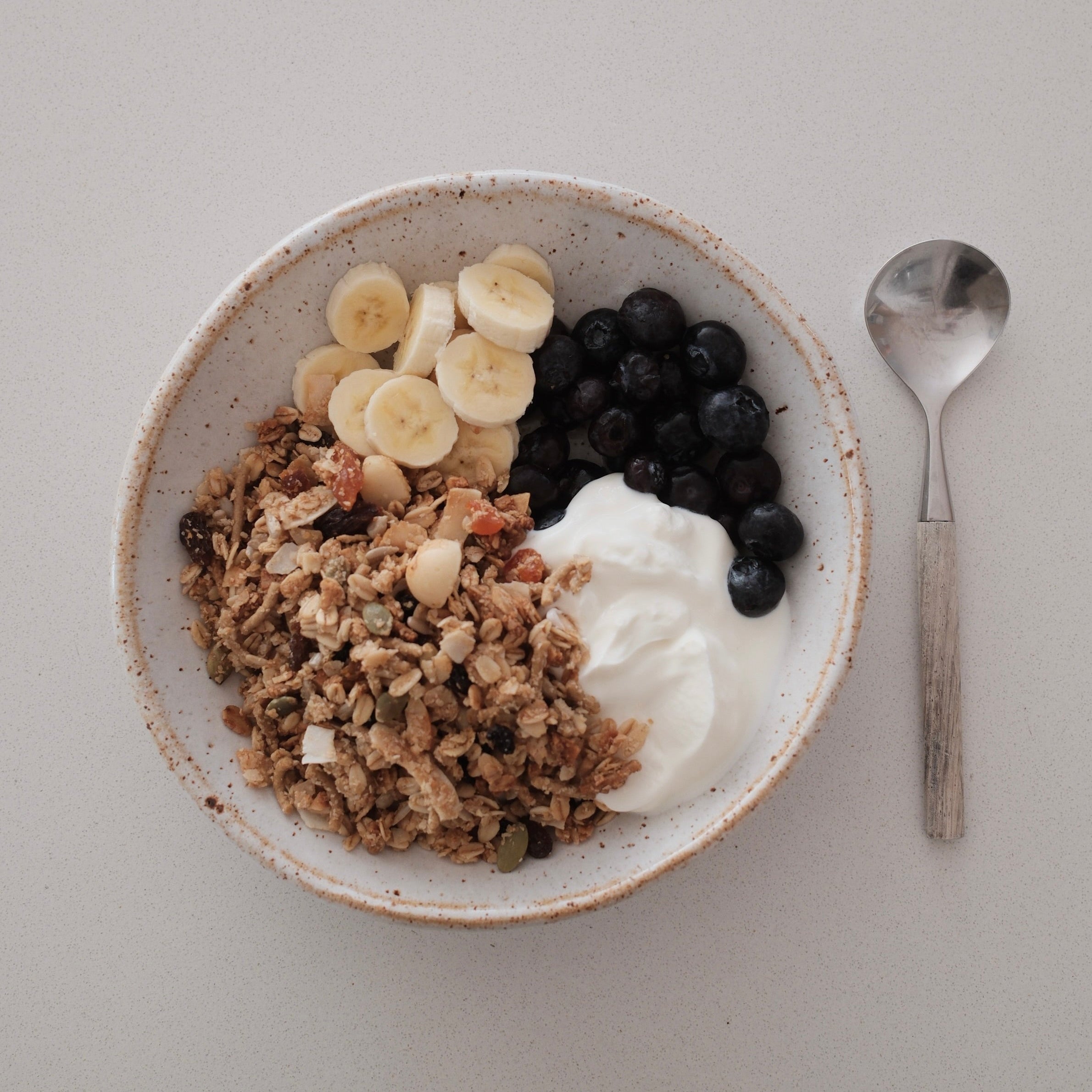 A top-down view of a white speckled ceramic bowl filled with granola, sliced bananas, blueberries, and a dollop of white yogurt, with a spoon placed to the right on a light surface.