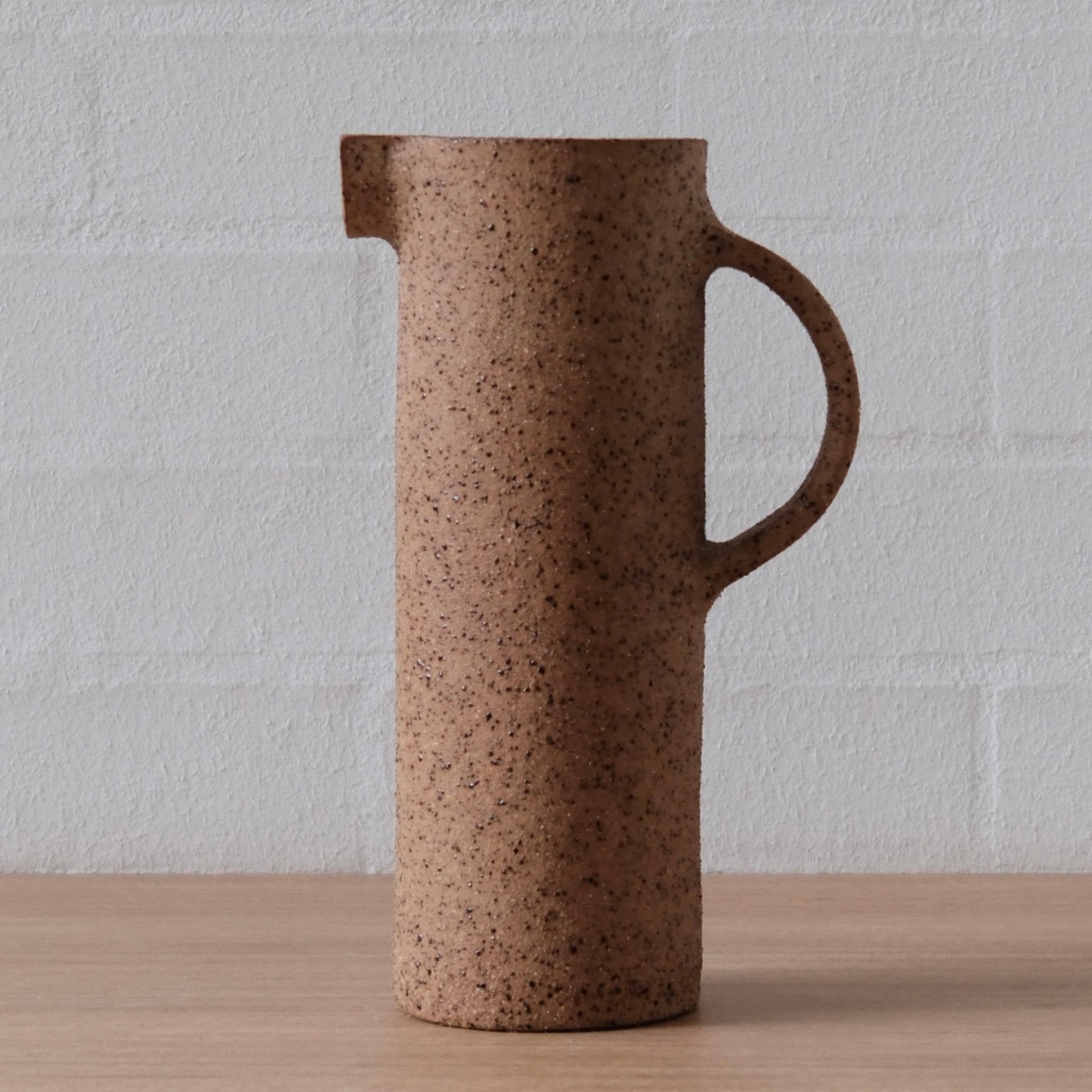 A speckled brown ceramic pitcher with a small pouring spout and a curved handle, standing on a light wood surface against a white brick wall.