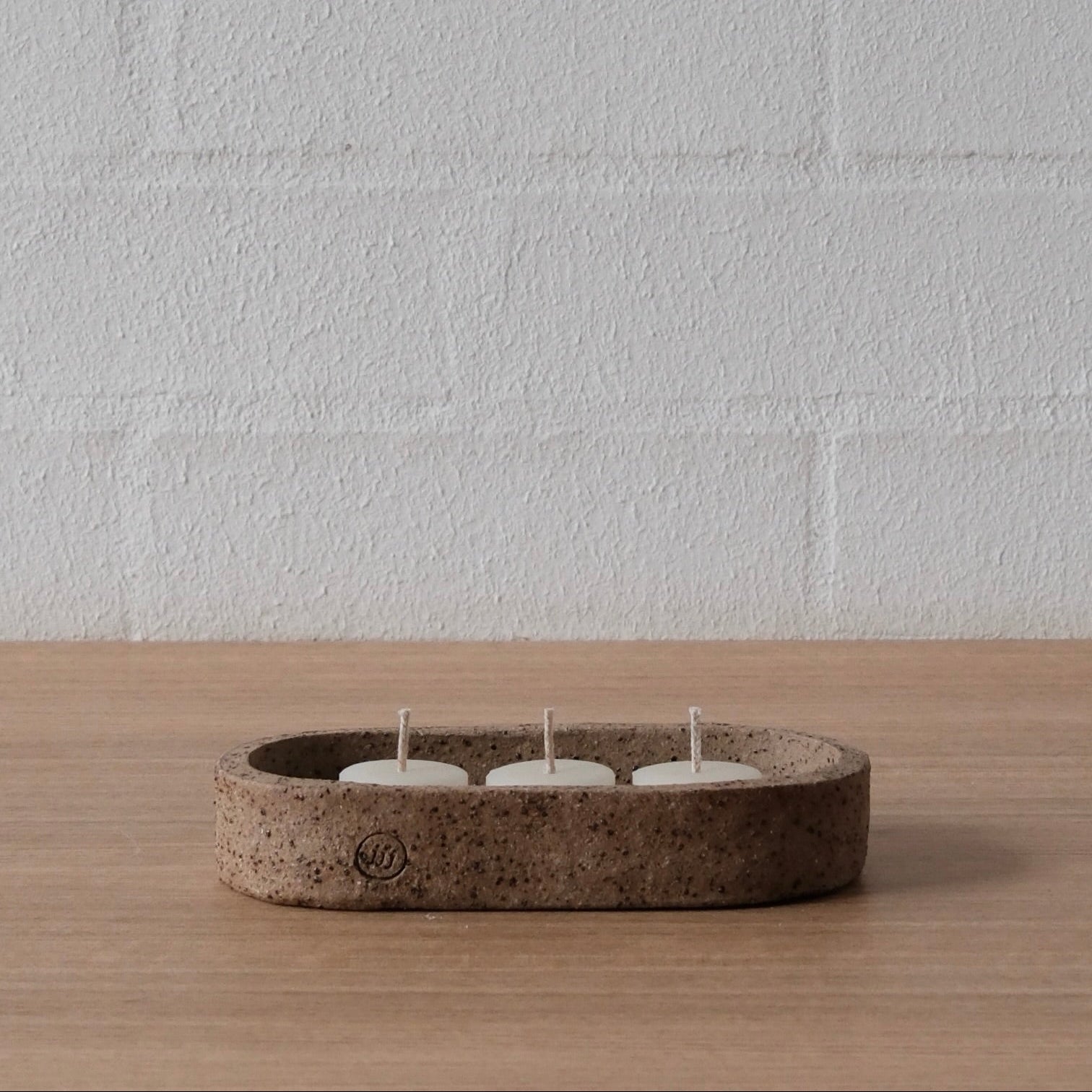 An oval-shaped tray, shallow dish round, or candle holder with three white tea light candles inside, set on a light wood surface against a white brick wall background.