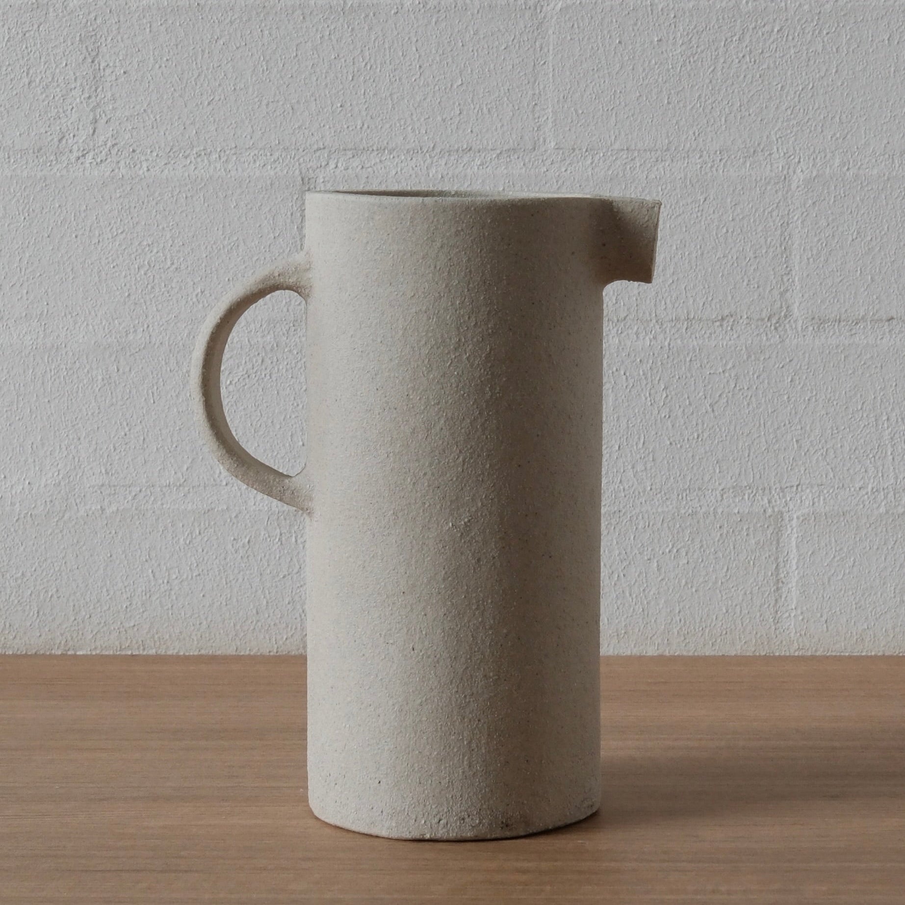 A white matte ceramic pitcher with a small handle and pouring spout, standing on a light wood surface against a white brick wall.