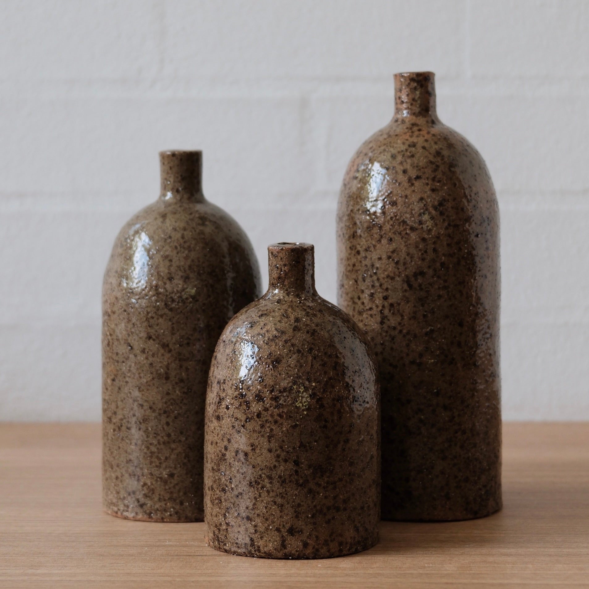 Trio of three olive speckled ceramic vases, with cylindrical body, rounded top and small spout, one a wooden bench in front of a white wall