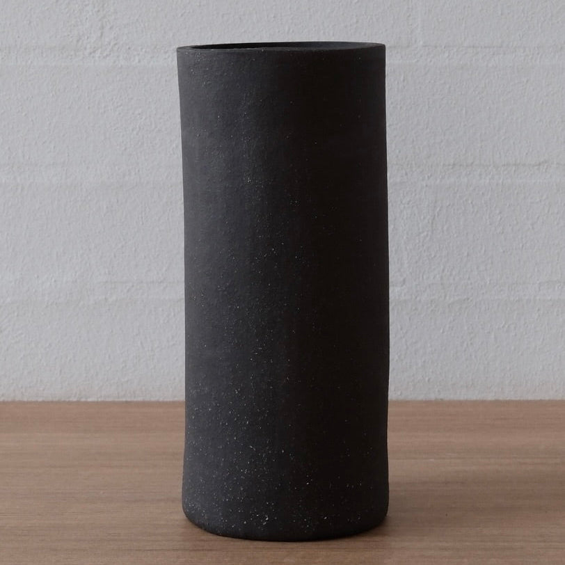 A matte black cylindrical ceramic vase with a textured, speckled finish, standing on a light wood surface against a white brick wall background.