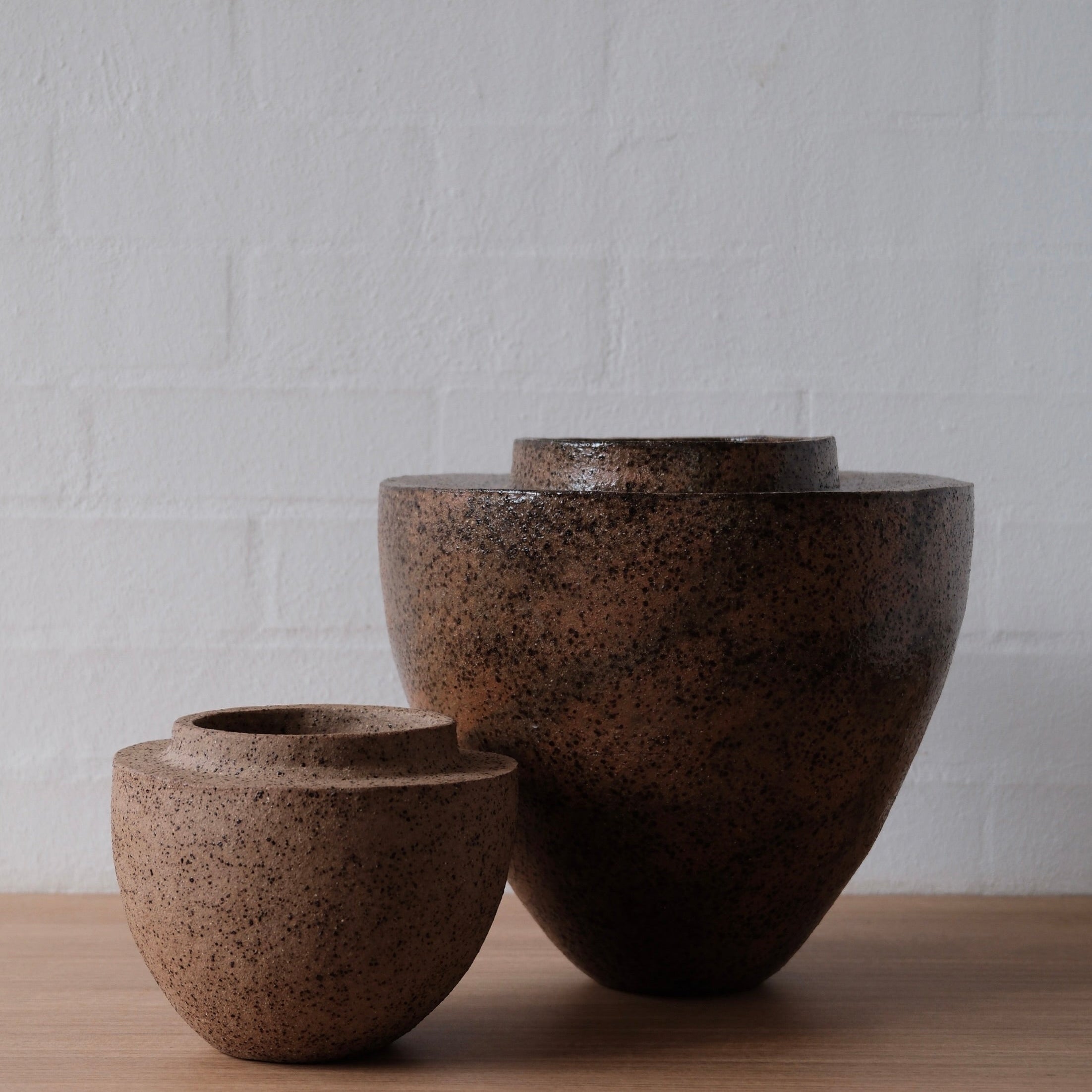 Two brown ceramic bowls, one small and the other large, on a wooden surface with a white textured wall background.