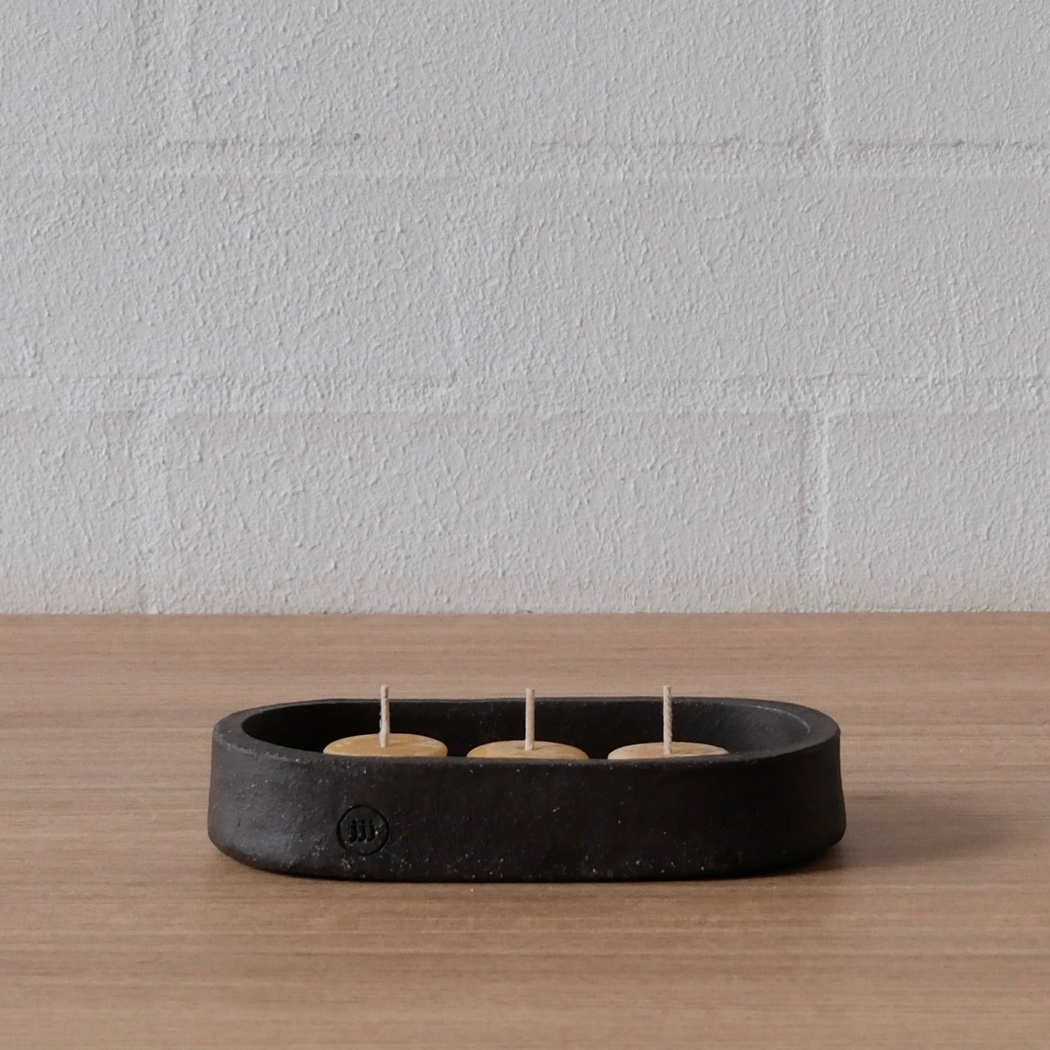 A black ceramic three-candle holder with three lit tealight candles, resting on a light wood surface against a white brick wall.
