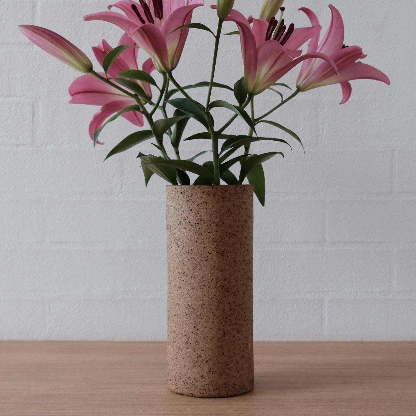 Pink lilies arranged in a tall cylindrical speckled brown ceramic vase, set on a light wood surface against a white painted brick wall.