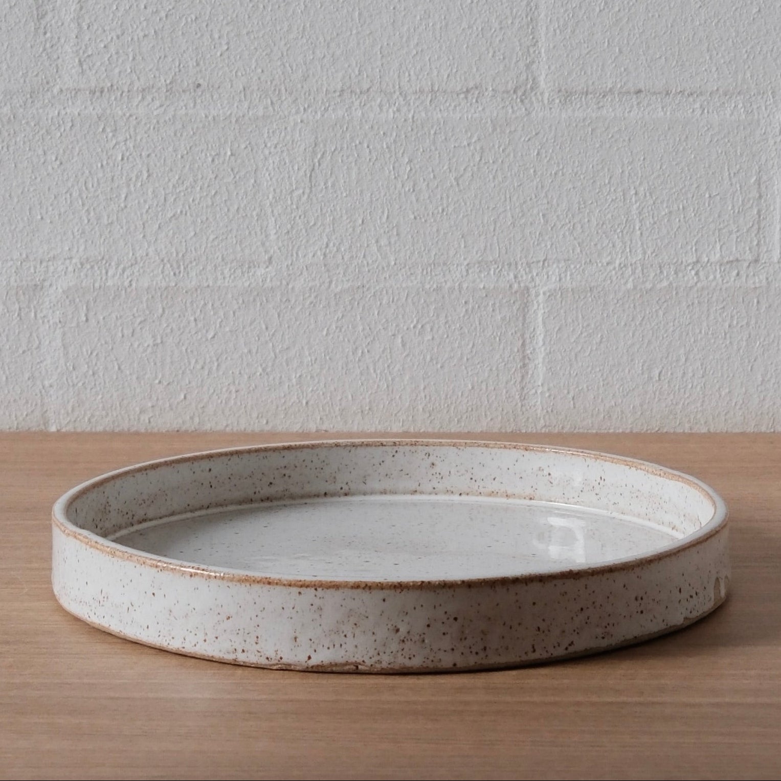 A large round ceramic tray with a speckled beige and white glaze, sitting on a light wood surface against a white brick wall background.