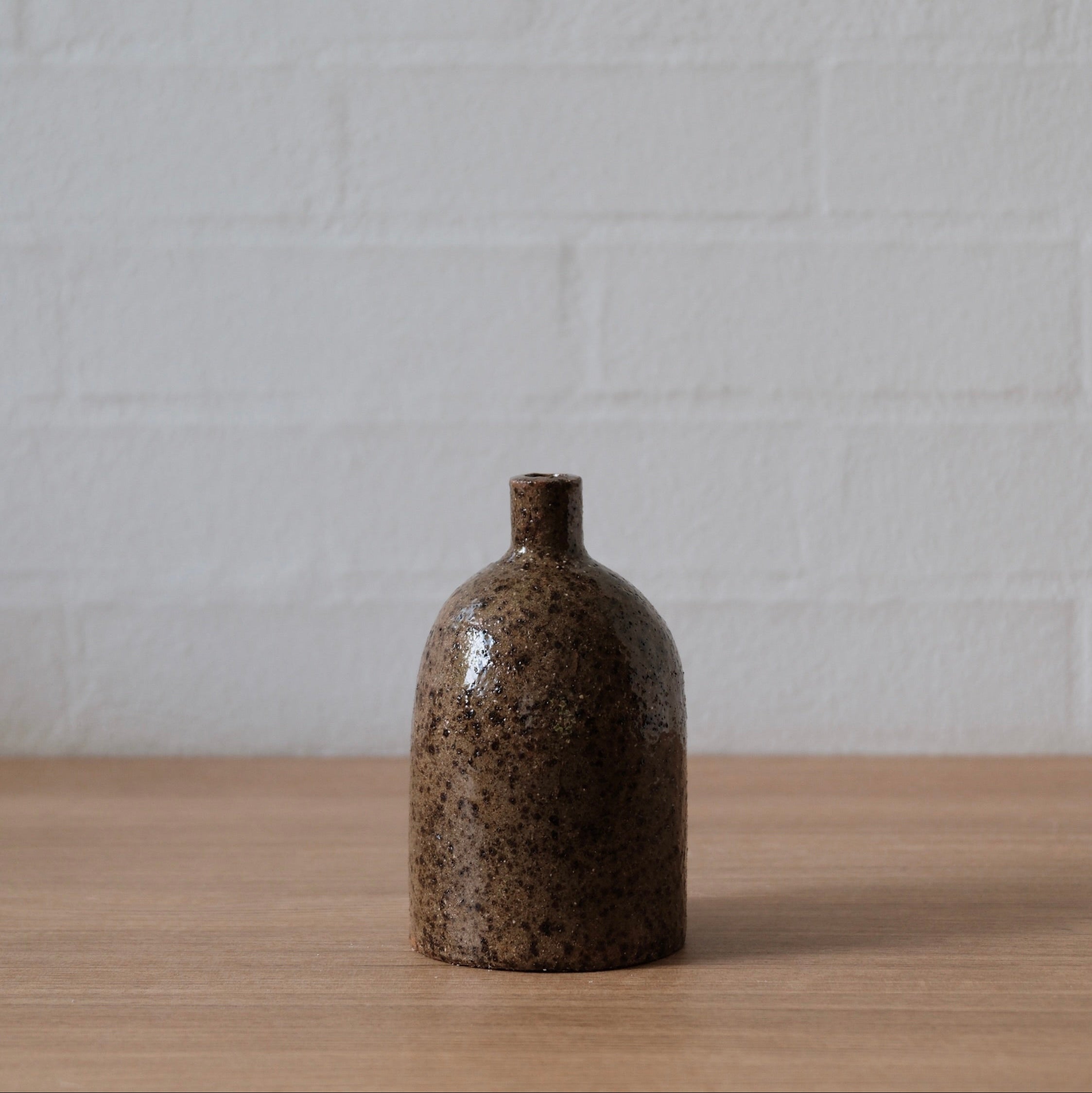 Small olive speckled ceramic vase, with cylindrical body, rounded top and small spout, one a wooden bench in front of a white wall