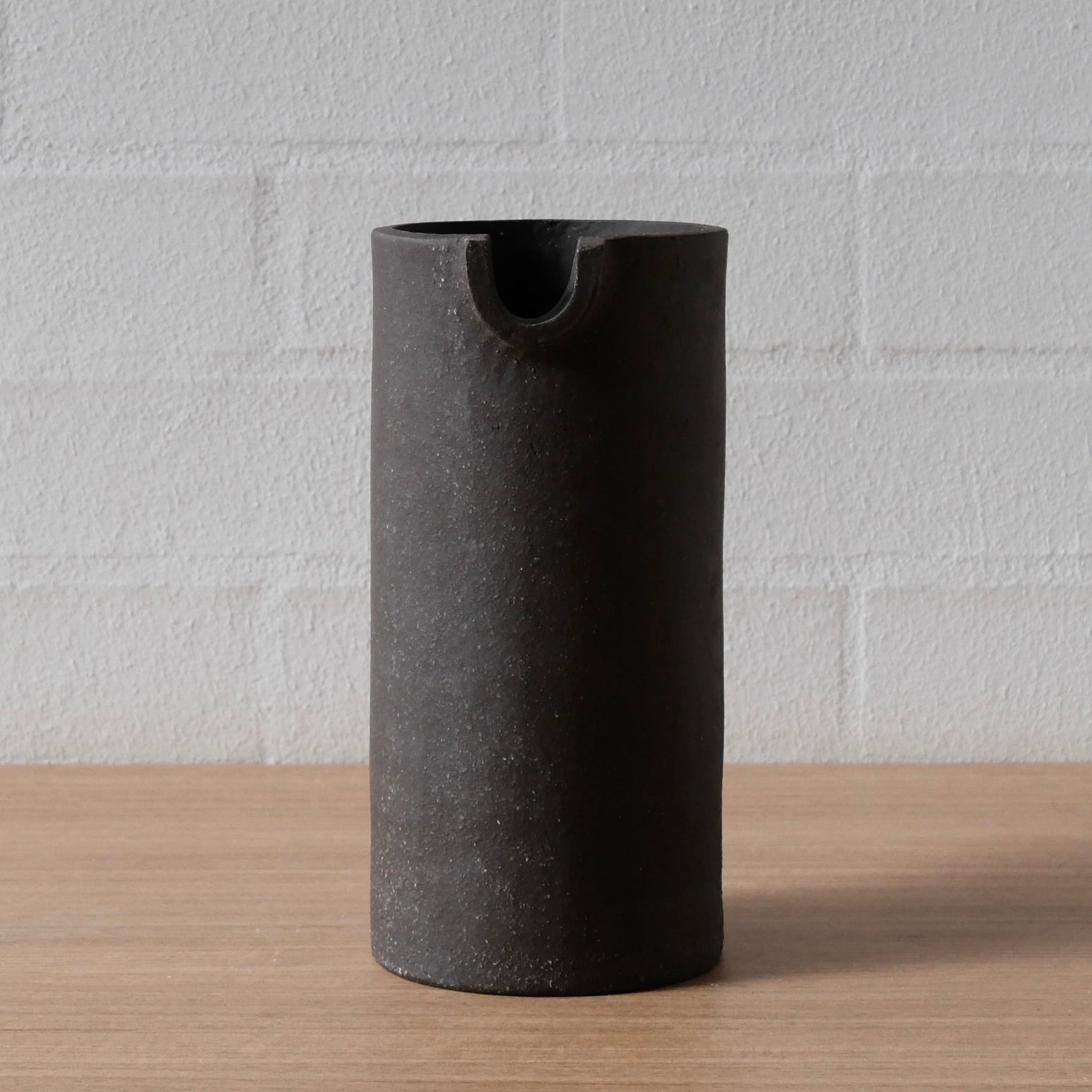 A matte black cylindrical vessel with a small pouring spout at the top, standing on a light wood surface against a white brick wall background.