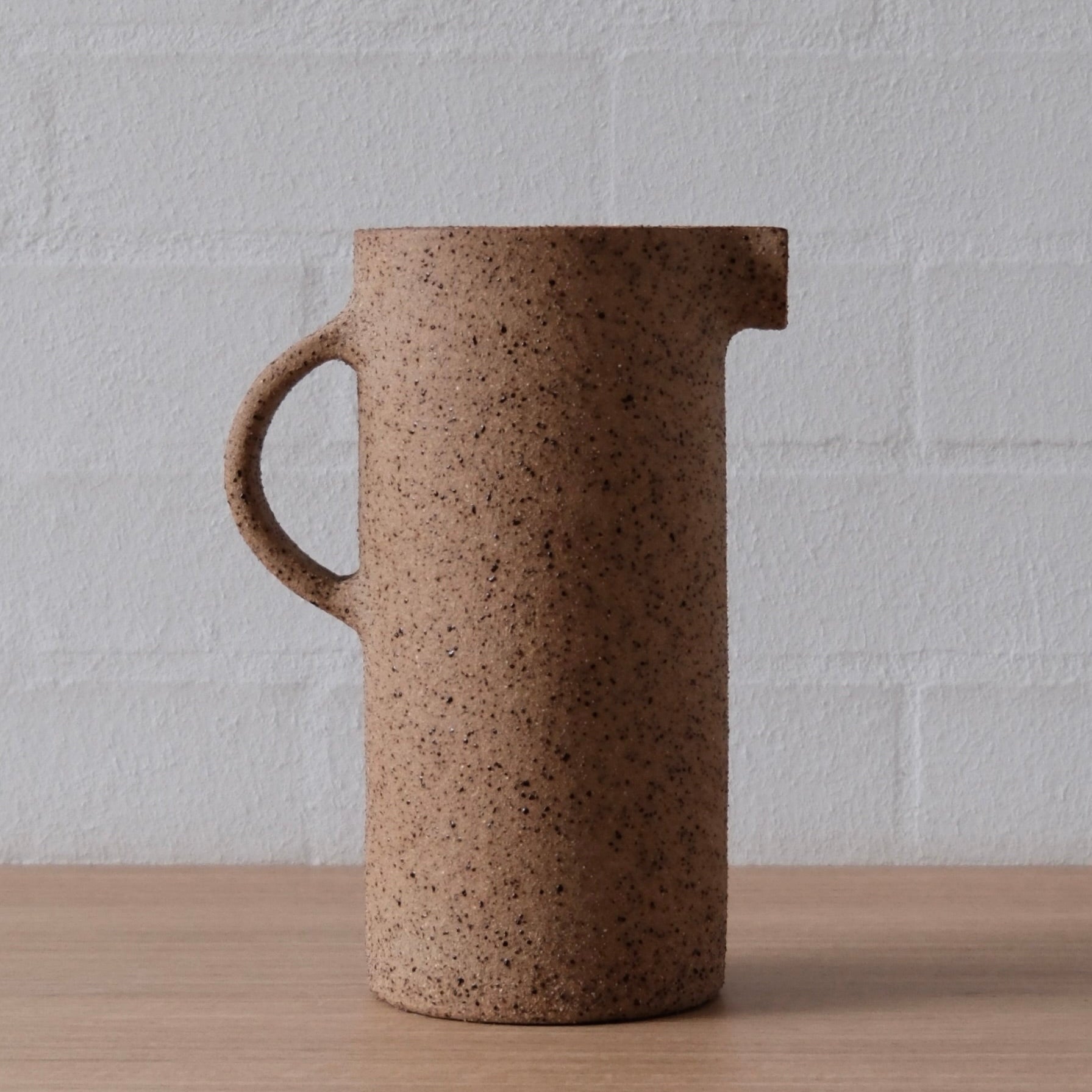 A speckled brown ceramic pitcher with a handle and pouring spout, standing on a light wood surface against a white brick wall background.