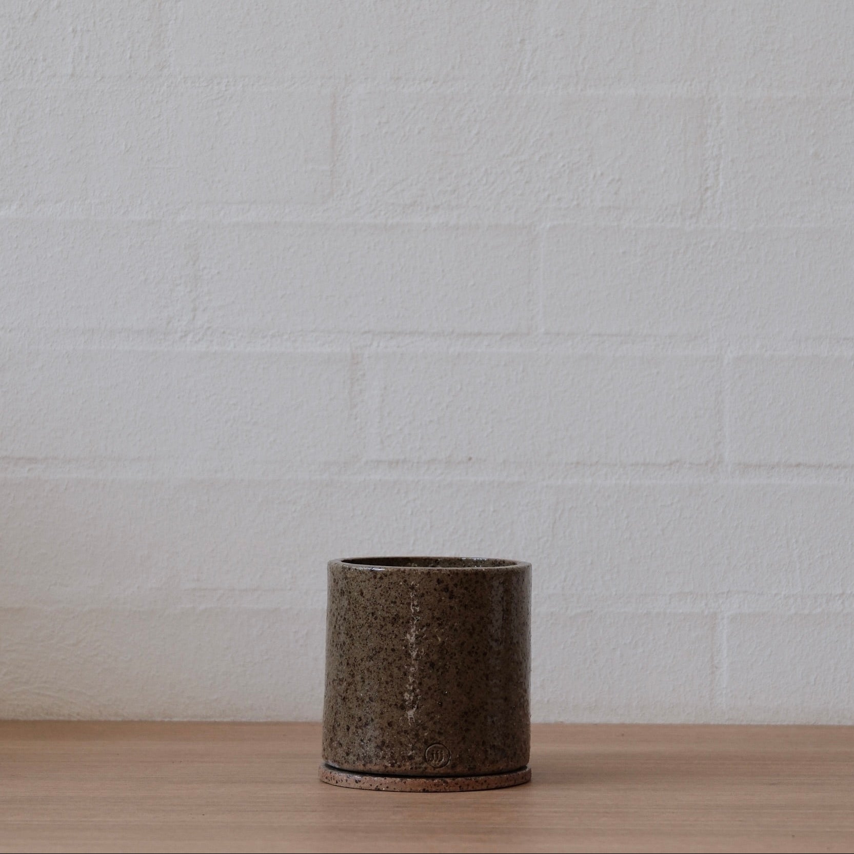 Small olive green with speckled ceramic pot on a wooden surface with a white brick wall background.