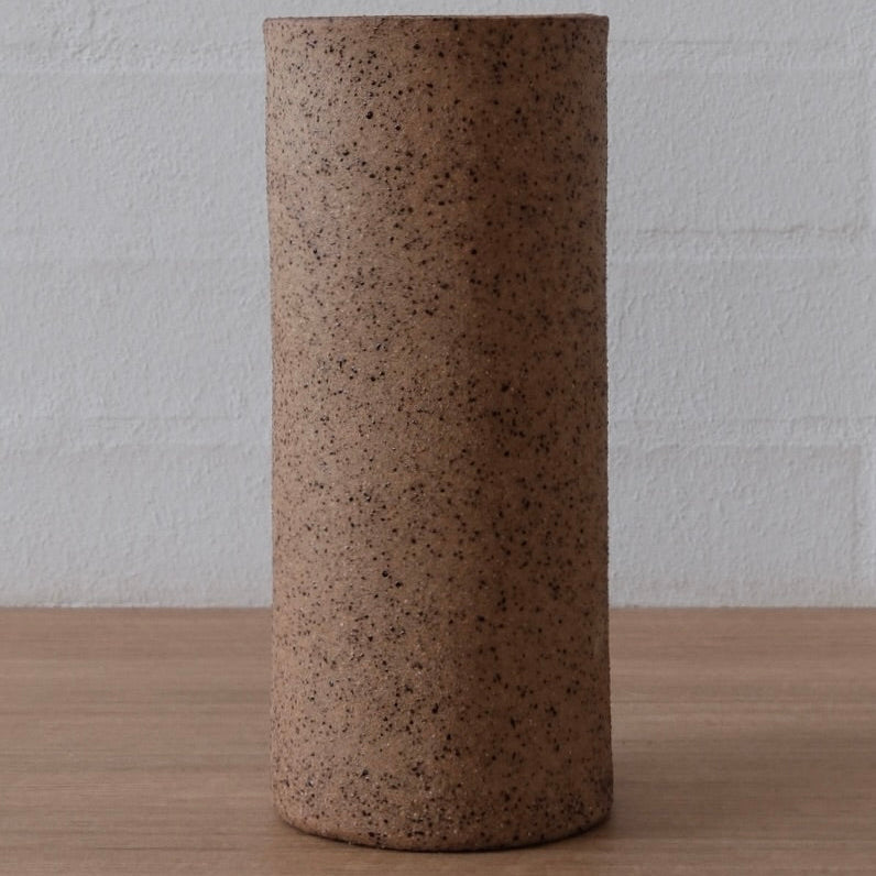 A cylindrical ceramic vase with a textured, speckled earthy brown finish, standing on a light wood surface against a white brick wall background.