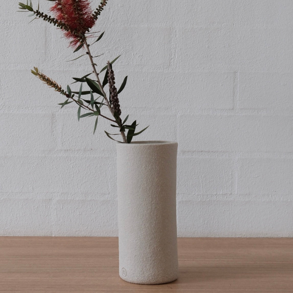 A tall, cylindrical matte white ceramic vase holding dried botanical stems with green leaves and a burgundy flower, set on a light wood surface against a white painted brick wall.