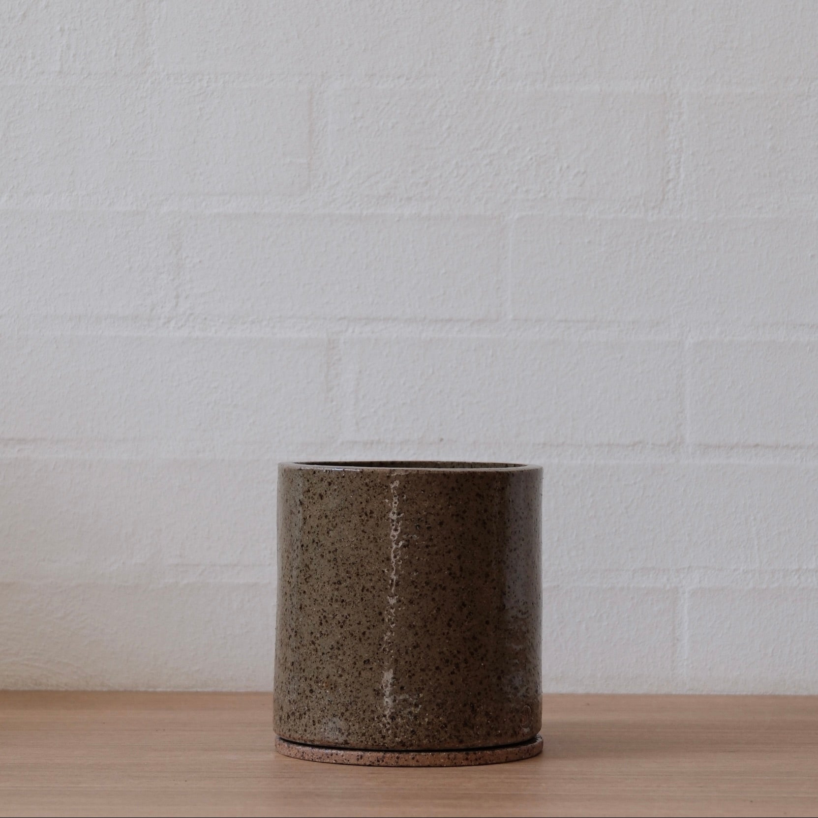 Medium cylindrical olive green with speckled ceramic pot on a wooden surface with a white brick wall background.