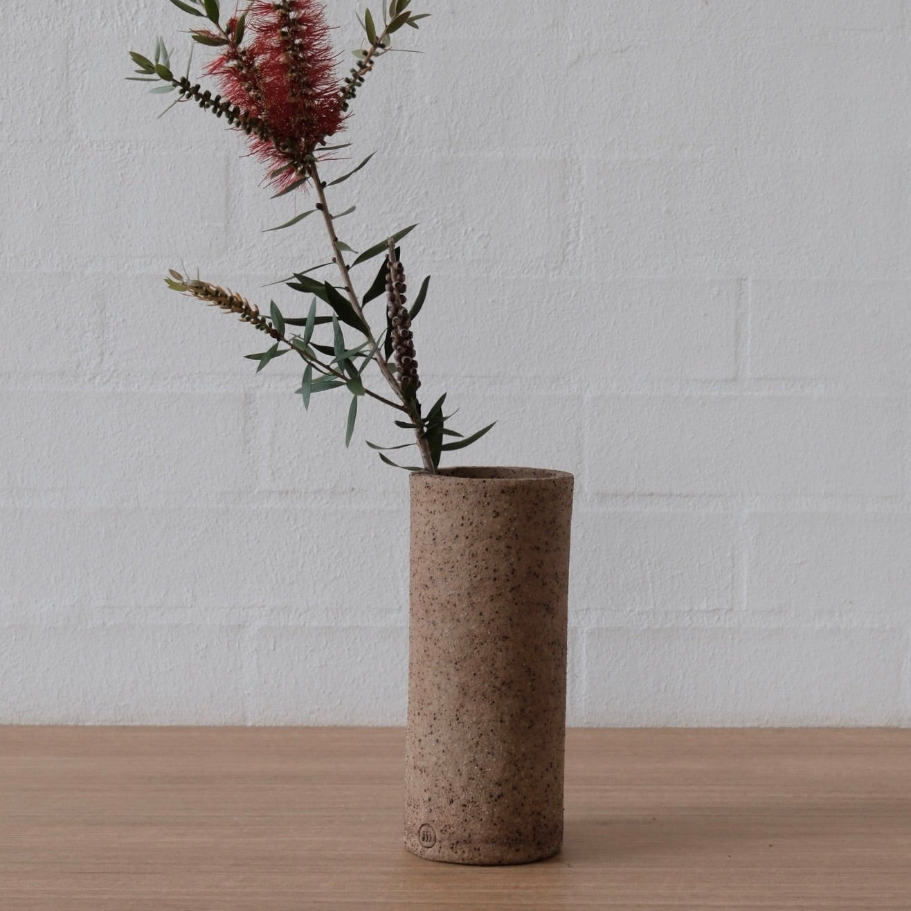 A medium cylindrical speckled ceramic vase in a warm brown tone, holding a single stem of pinkish-red bottlebrush flowers with green leaves, set on a light wood surface against a white painted brick wall.