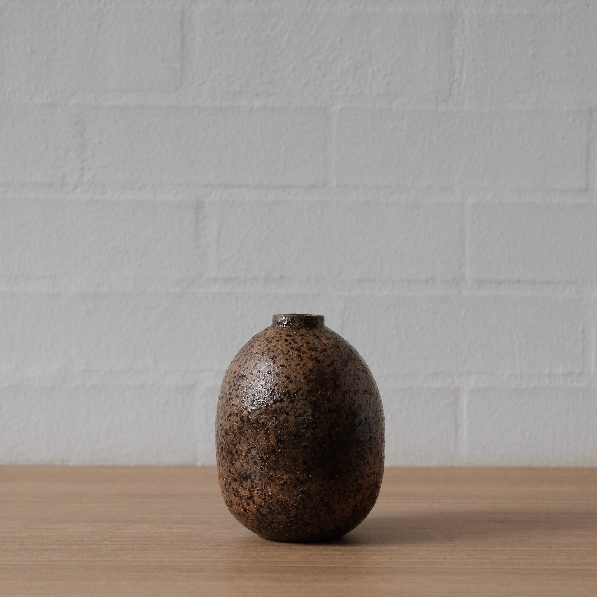 Brown speckled ceramic vessel on a wooden surface with a white textured wall background
