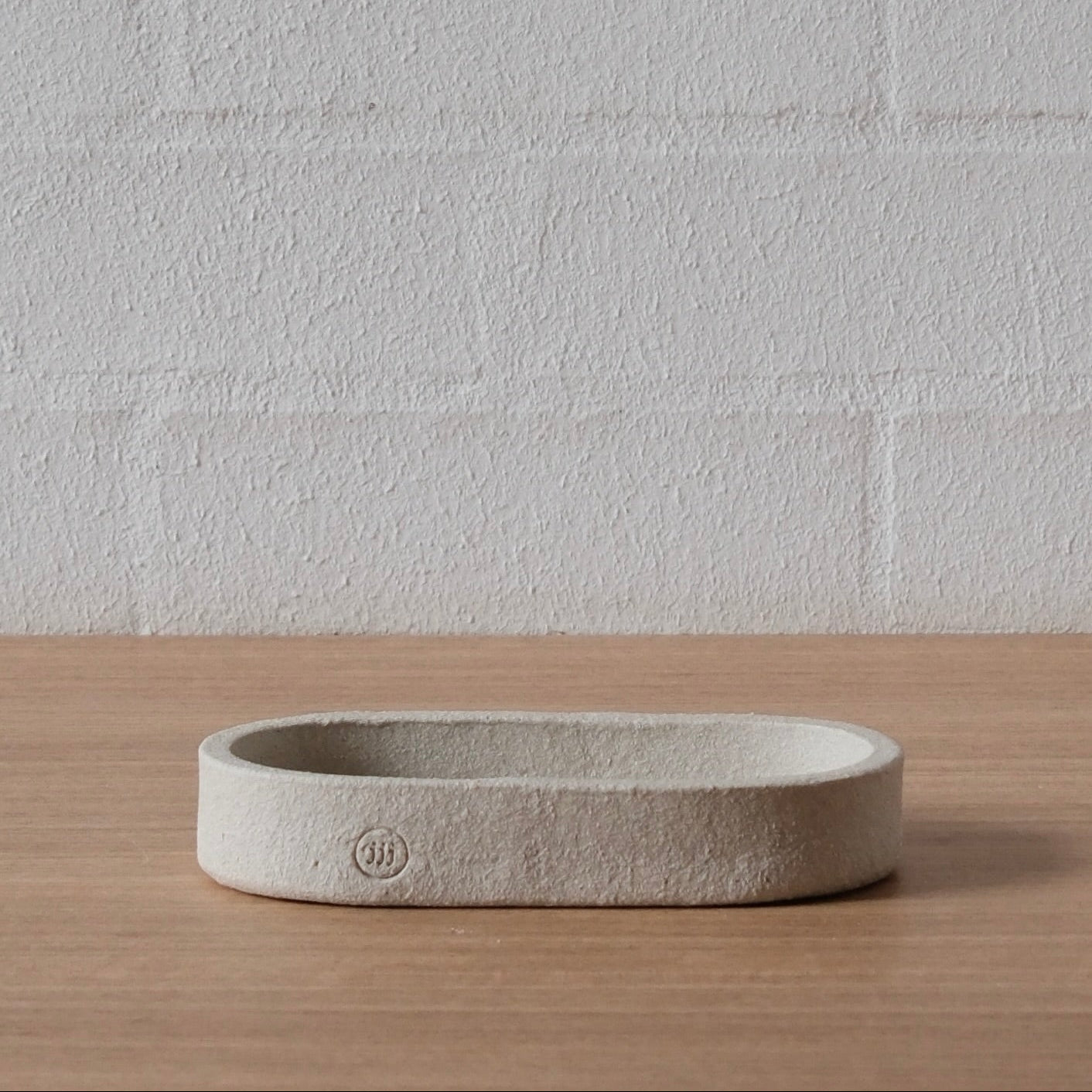 A minimalist, low-profile oval tray or shallow dish in a textured white ceramics, resting on a light wood surface against a white brick wall background.